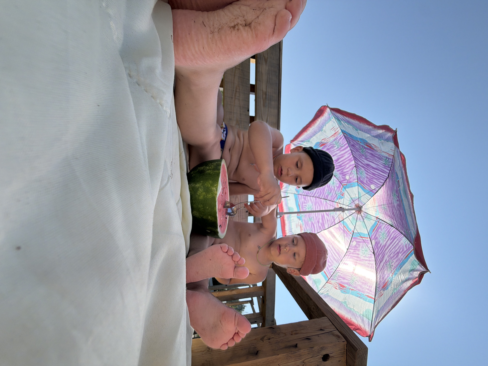 In Chaikovsky, Russia, two young boys are seated on a white blanket under a colorful umbrella, sharing a halved watermelon. The boy on the left, wearing a black beanie, is using a spoon to scoop watermelon. The boy on the right, wearing a reddish beanie, is observing. Both boys are shirtless. Their feet are visible in the foreground, with the boy on the left's left foot showing dirt. They are positioned on a wooden bench, with a wooden railing to the right. The sky above is clear blue. The image is captured from a low angle, looking upwards.