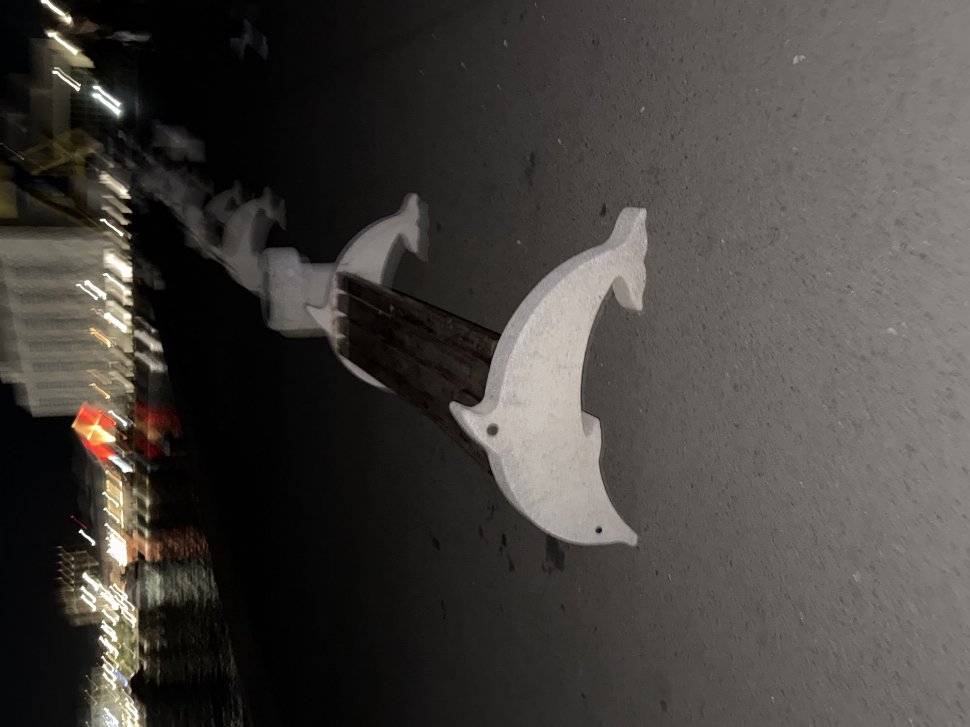 The image depicts a bollard at night, likely in Burgas, Bulgaria, given the dolphin-shaped design. The bollard is composed of a dark, horizontal wooden beam supported by three white, stylized dolphin-shaped brackets. These brackets are spaced along the beam. The bollard is placed on a gray concrete or paved surface. In the background, blurry lights from buildings are visible, suggesting an urban setting at night. The focus is on the bollard, with the background providing context.