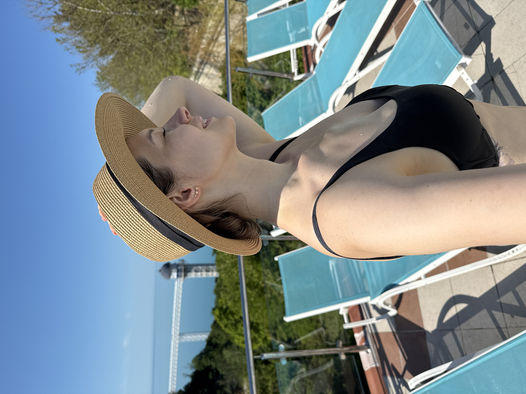 An adult female is depicted in profile, facing right (viewer's left), on an outdoor terrace in Nebug, Russia. She wears a straw hat with a black band and a black bikini top. Her left arm is raised, with her hand resting on or near the hat. Her eyes appear closed or downcast, and she exhibits a slight smile. A three-star stud earring is visible in her left ear. The terrace features a light-colored tiled surface and multiple turquoise-colored lounge chairs with white frames. A railing with metal posts and glass panels separates the terrace from the background. Beyond the railing, dense green foliage is visible, descending towards a large body of water identified as the Black Sea. In the mid-distance over the water, a white bridge-like structure with a dark-domed tower (likely an observation tower) is prominent. The sky is clear and blue, indicating a sunny day.
