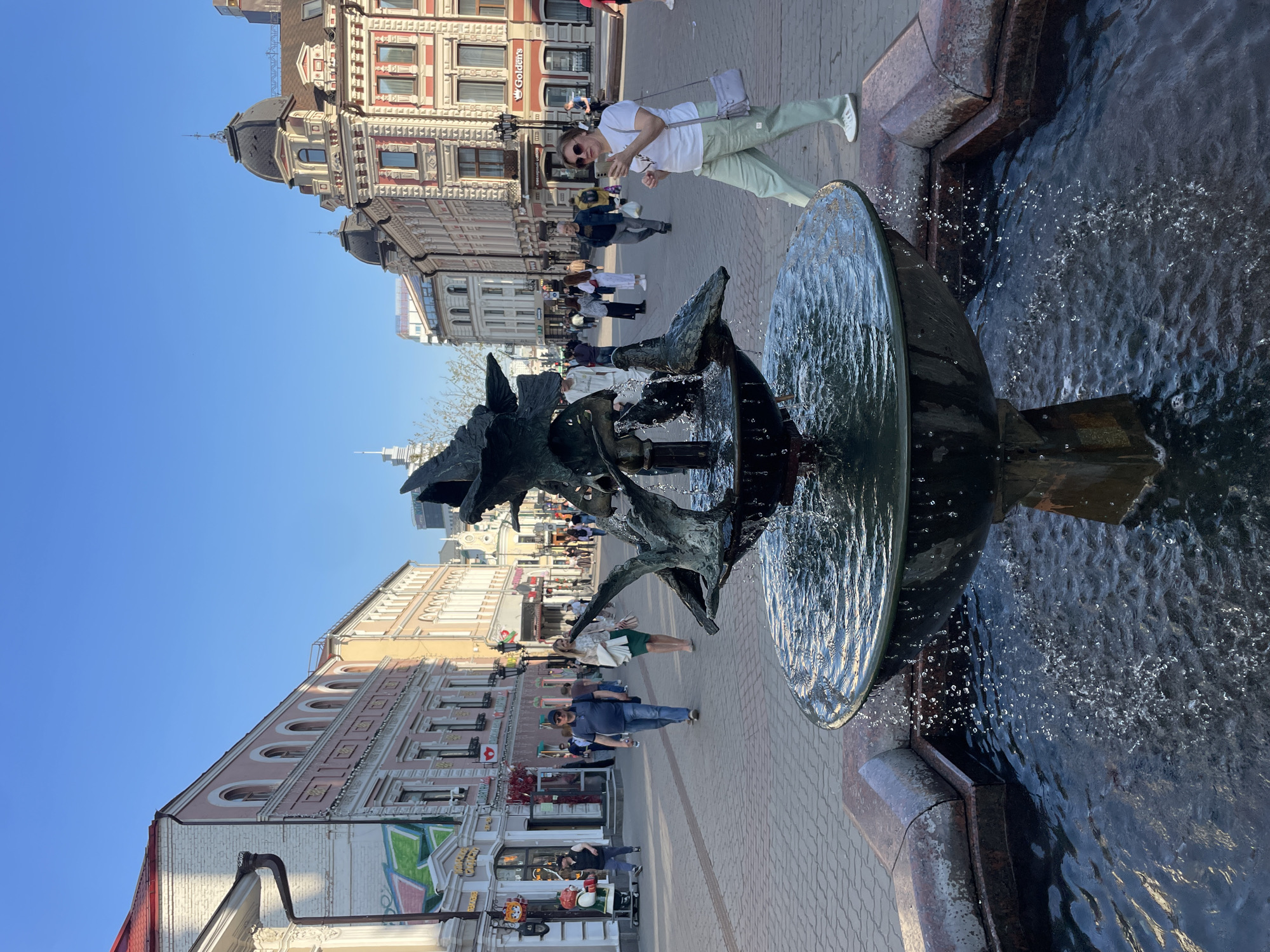 The image captures a view of Bauman Street in Kazan, Russia. A multi-tiered fountain with a black metal sculpture of what appears to be dragon-like figures is in the foreground. Water is actively flowing from the fountain into a pool.
In the background, a brick-paved pedestrian street is lined with multi-story buildings of varying architectural styles, some with intricate facades and domes. People are walking along the street. A woman in a white shirt and light green pants is standing near the fountain. She is looking at her phone and seems to be taking a photo. The sky is clear and blue, suggesting a sunny day.