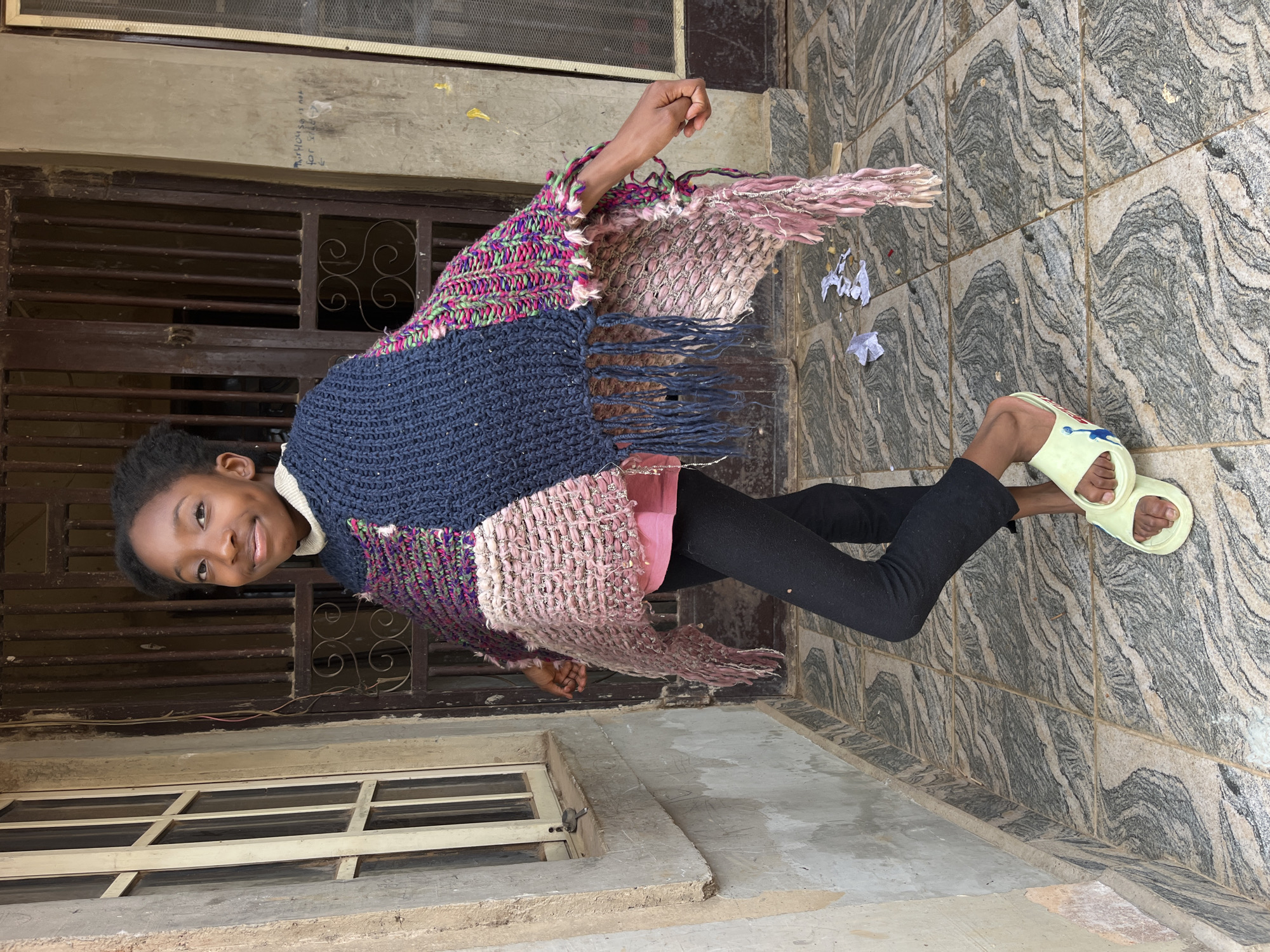A young Black girl stands outside, striking a pose. She wears a multicolored knitted poncho over a pink shirt and black leggings, completing her outfit with light yellow slides. The girl stands on a tiled patio in front of a building with a window and a closed metal door. The background is a wall with faded graffiti. The overall setting is that of an outdoor urban environment.