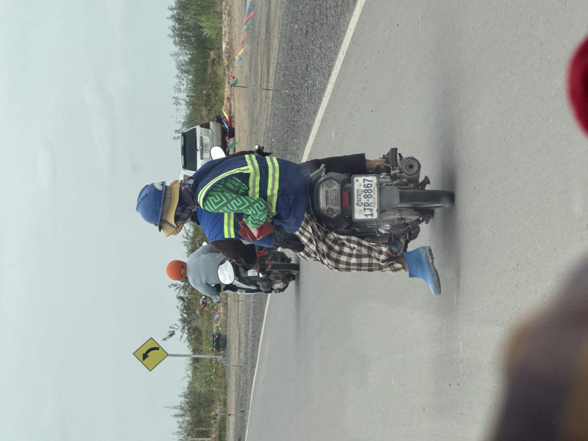 The image shows two people on a motorcycle, with the rider in front wearing an orange helmet and a light gray jacket, and the passenger behind wearing a blue helmet, a yellow hat, and a green and white patterned vest over a blue jacket. The passenger is also wearing plaid pants and blue boots. The motorcycle has a license plate that reads "1JR-8867" and "PHNOM PENH". They are on a paved road, likely in or near Krong Ta Khmau, Cambodia, given the license plate information. In the background, there is a white SUV, a yellow directional sign indicating a curve to the left, and various flags or banners strung along the side of the road. The environment appears to be a roadside with some vegetation and construction or unpaved areas visible. The sky is overcast.