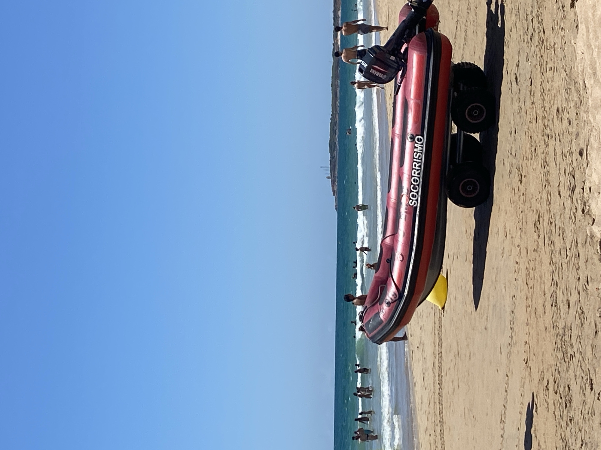 An inflatable rescue boat with the text "SOCORRISMO" is on the sandy beach in Conil de la Frontera, Spain. The boat is red with black trim and is mounted on a trailer with four wheels. A Yamaha outboard motor is attached to the stern of the boat. The sea is calm with gentle waves and a clear blue sky overhead. Several people are in the water, swimming and playing in the waves. In the background, a peninsula with some structures is visible in the distance. The scene depicts a typical summer day at the beach.