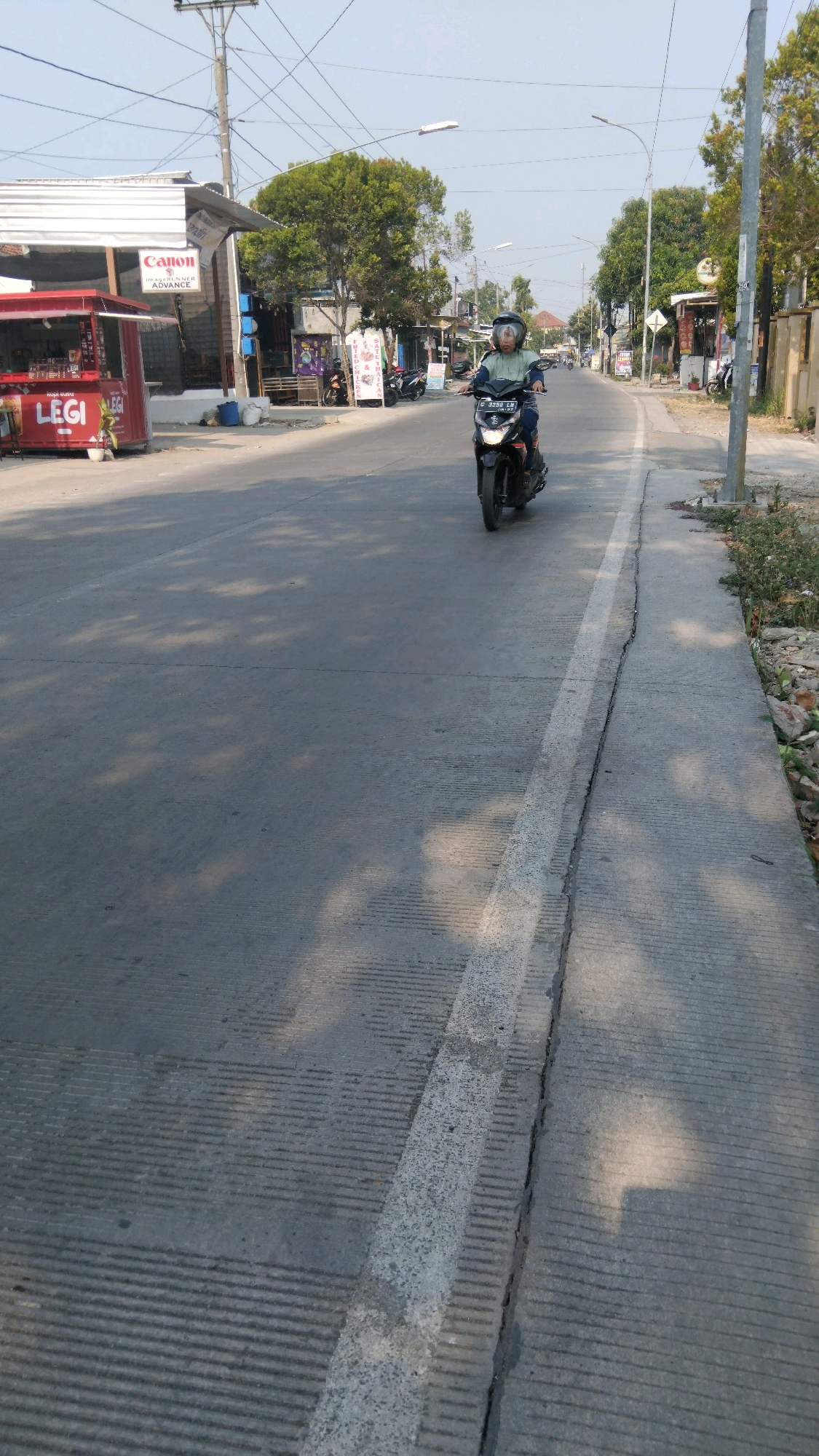 A person wearing a green jacket and a helmet is riding a dark-colored motorcycle on a road in Tegal, Indonesia. The road has a concrete surface with textured lines and a white line marking the lane. Sunlight casts shadows on the road from trees and overhead wires. On the left side of the road, there are several small businesses, including one with a red canopy and signs that say "Canon" and "LEG I". On the right side of the road, there is a sidewalk with some vegetation and rocks. The sky is clear and blue.