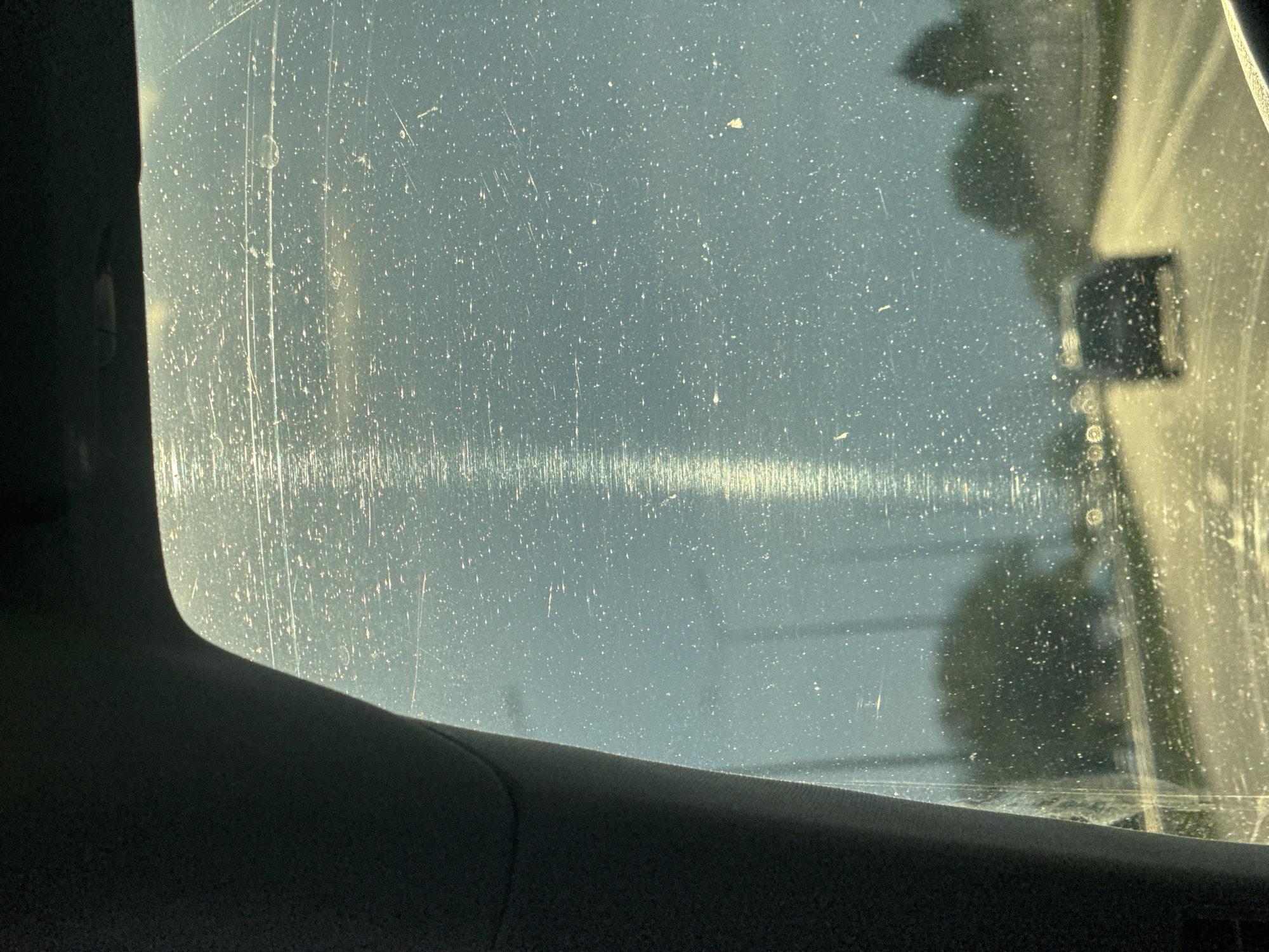 The image captures an exterior scene in Suprunivka, Ukraine, viewed through a heavily soiled and scratched vehicle windshield. The windshield is uniformly covered with numerous small white specks, indicative of dust or water spots, and crisscrossed by multiple fine scratches. A prominent vertical streak of intense light, consistent with sun glare, extends from the top of the windshield to the middle, illuminating a path through the debris.
The interior of the vehicle is visible on the left and top, featuring dark grey fabric trim, likely part of an A-pillar and headliner.
Beyond the obstructed glass, a paved road stretches into the distance, reflecting a yellowish light, suggesting a low sun angle. A dark-colored vehicle, appearing as a blurred silhouette, is observed moving away from the viewer in the middle distance on the right side of the road. Additional indistinct light sources, possibly streetlights or other vehicles, are visible further down the road on the left. Blurred forms of trees and structures line both sides of the road, appearing dark against a hazy, bluish-grey sky. No human figures are discernible.