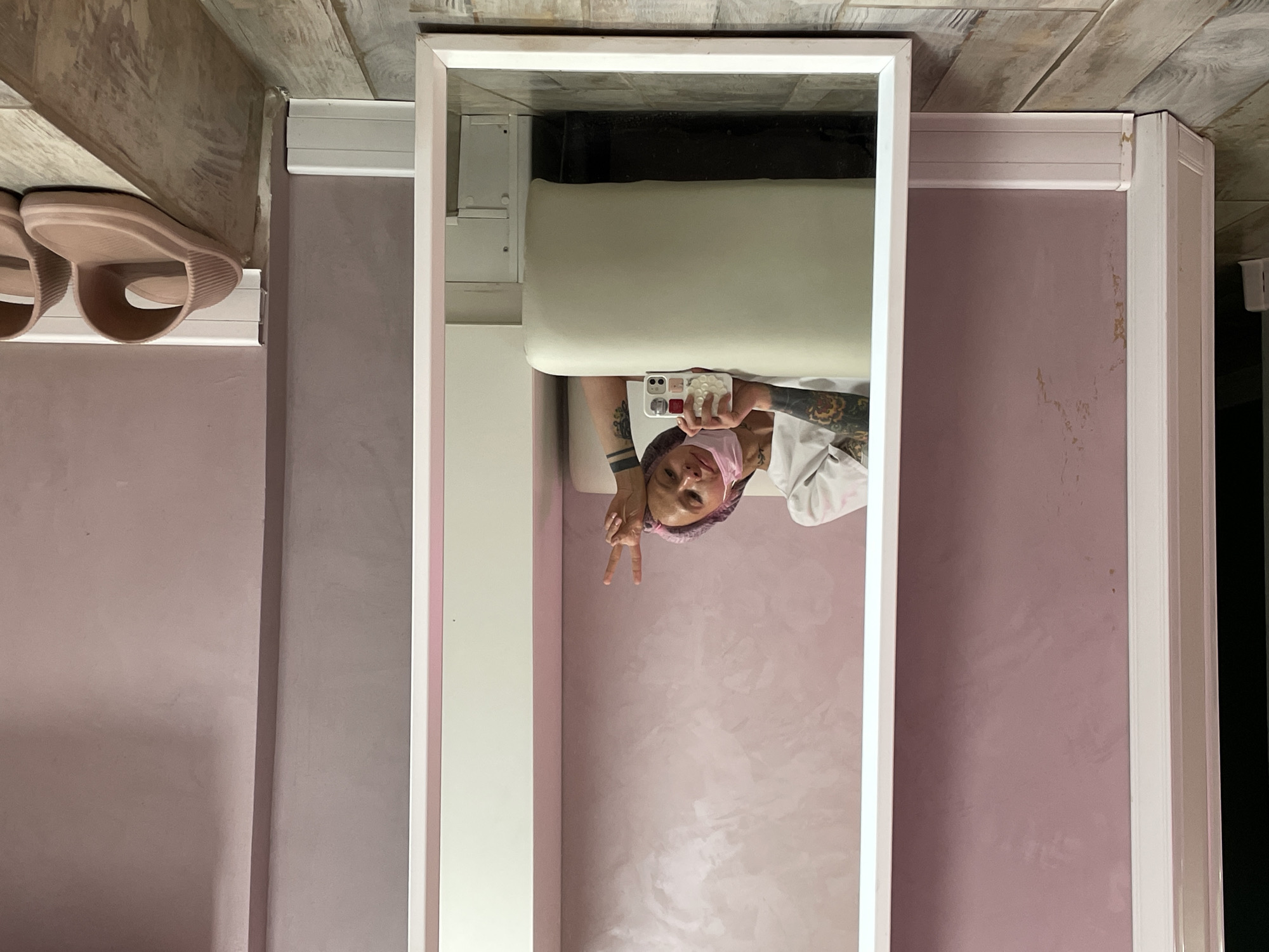 The image shows a vertically oriented view of a bathroom interior, likely in Izhevsk, Russia. A woman is visible in the mirror's reflection, holding a phone up to take a selfie. She is wearing a face mask and head covering, and has visible tattoos on her arm. She is giving a peace sign with her fingers. Behind her, the reflection reveals a white examination table or bed and a small, unidentified apparatus mounted on the wall. The wall is painted a textured shade of light purple. The bathroom is decorated in light shades. A pair of pink slides are visible on a shelf in the upper left corner of the frame. The ceiling is made of horizontal wooden planks.
