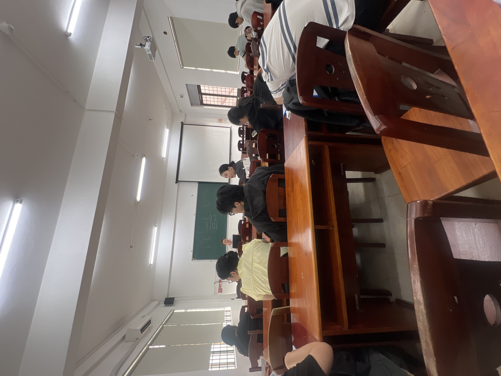 The image, rotated 90 degrees clockwise, captures a classroom scene in Đà Nẵng, Vietnam. The perspective is from the front-right of the room, looking towards the back and left side.
**Subjects:** Numerous students, appearing to be young adults or teenagers, are seated in rows at dark brown wooden desks and chairs. Most are visible from behind or in side profile, facing the front of the classroom. They wear casual clothing, including t-shirts and long-sleeved shirts. The classroom features white walls and a white ceiling with multiple illuminated fluorescent light fixtures. On the back wall, there is a green chalkboard with white writing and a blank white projection screen. A ceiling-mounted projector is visible near the front. Windows on the left wall are partially covered by blinds. Air conditioning units and electrical conduits are also visible on the walls and ceiling, along with a fire alarm/smoke detector.
**Activity:** The students are engaged in a learning activity, likely a class or study session. Their postures suggest focus, with many looking down at their desks, presumably reading or writing, while some observe the chalkboard or projection screen.
**Notable Details:** The interior is brightly lit by artificial lights. The presence of window blinds indicates it could be daytime, but inside. The wooden furniture and classroom layout are characteristic of educational settings. Students appear calm and focused.
**Visible Text:** The green chalkboard displays white writing, which appears to be mathematical or scientific notations and symbols. A red and white sign on the wall clearly reads "PCCC 23 CỨU HỘ," which translates from Vietnamese to "Fire Prevention and Fighting, Rescue 23."