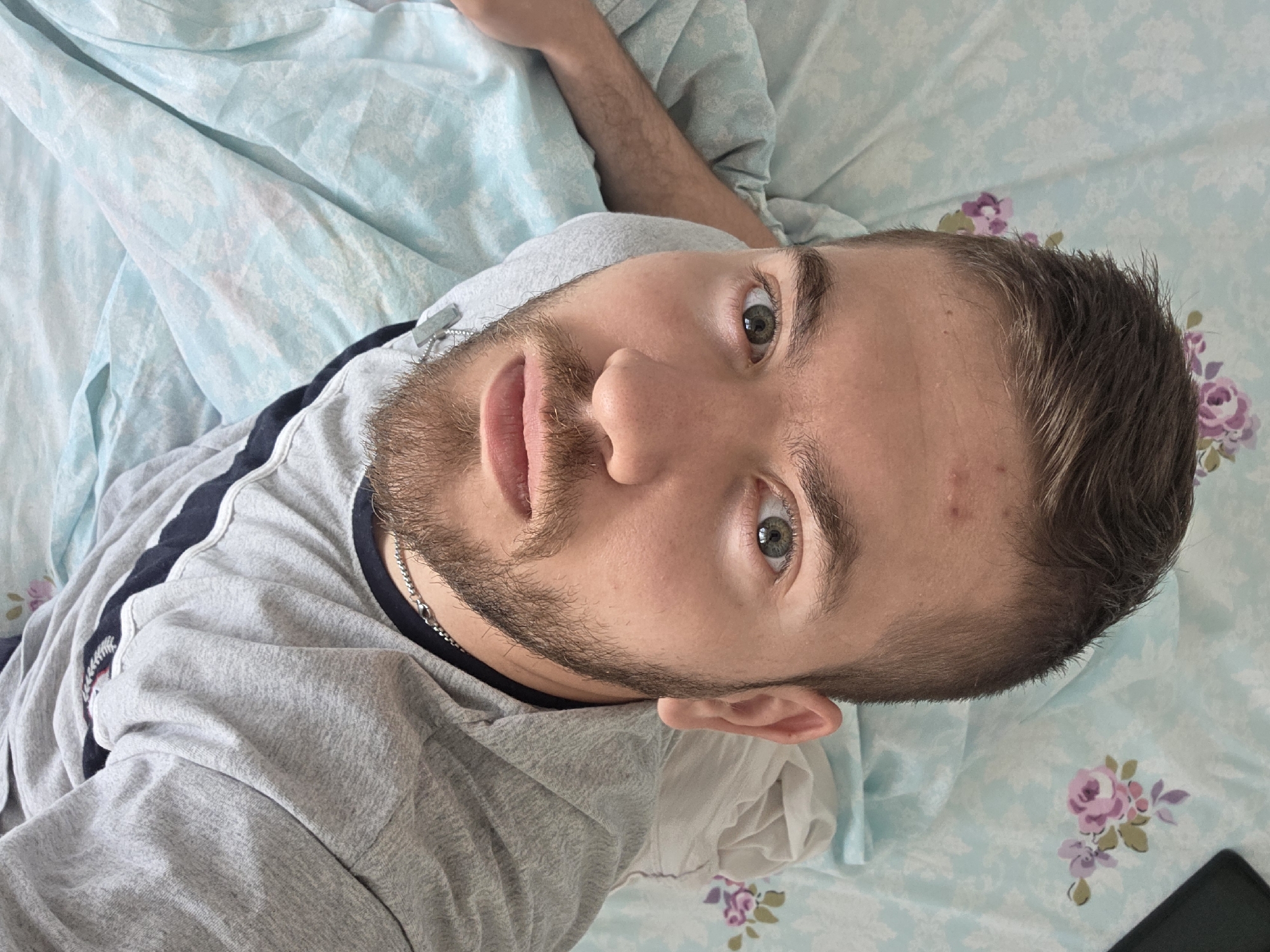 A close-up, upward-angled photograph captures a fair-skinned male with short brown hair and a trimmed beard. He lies on a light blue floral-patterned bed sheet. The man is wearing a grey t-shirt with a dark blue stripe down the side. A silver chain is visible around his neck. His gaze is directed towards the camera. The background is out of focus and appears to be a bed covered with patterned bed linens. The location in the city of Yagodovo, Bulgaria cannot be determined from the visual information.
