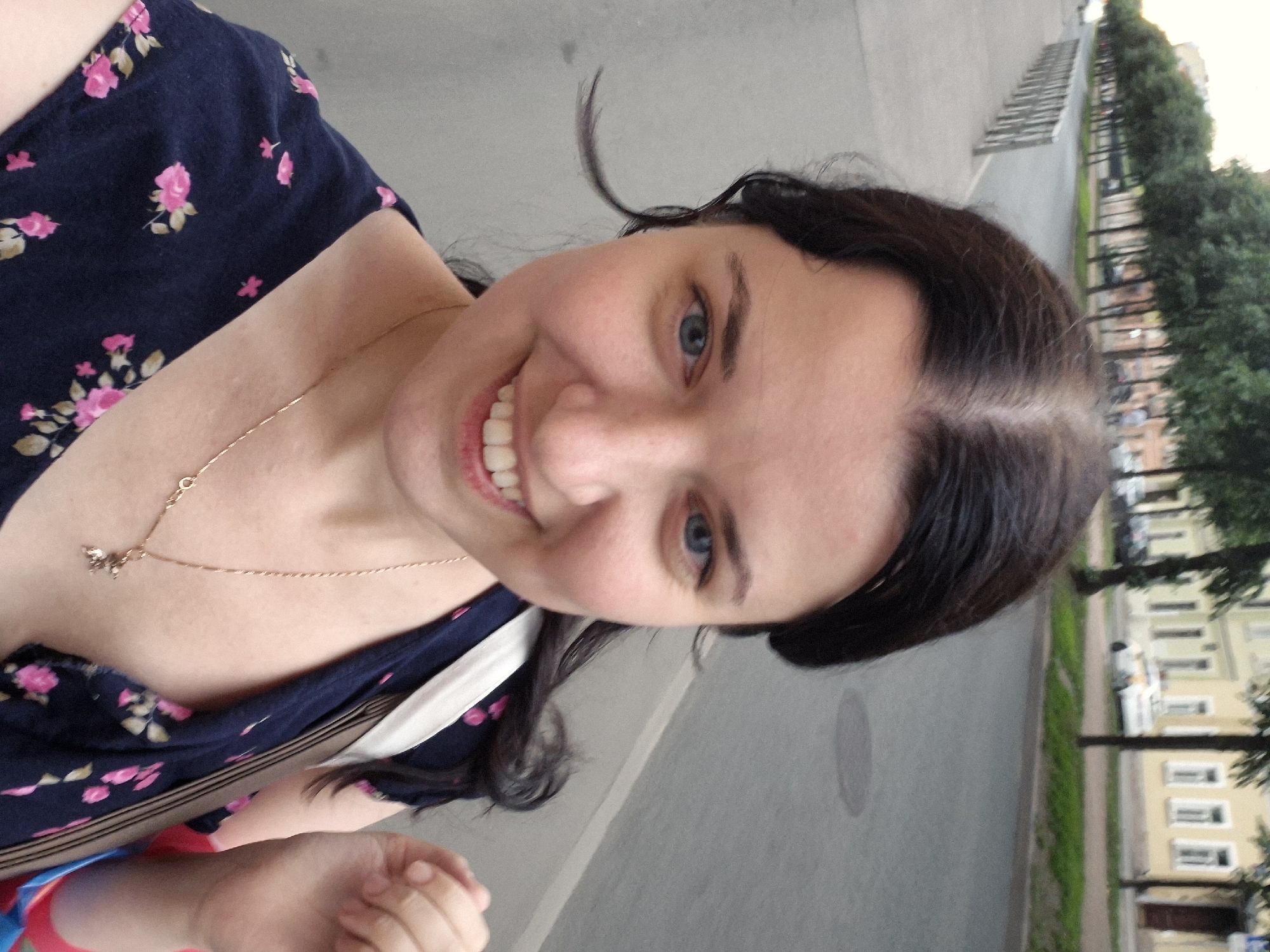 This is a selfie taken outdoors in Saint Petersburg, Russia. The primary subject is a young woman with dark hair and blue eyes, smiling widely and showing her teeth. She is wearing a dark blue dress with a floral pattern of pink roses. A delicate gold chain with a small charm rests on her collarbone. Her left arm and hand are visible in the lower part of the frame.
The background suggests a street scene with a sidewalk lined with trees and buildings. The architecture of the buildings, with their light-colored facades and numerous windows, is characteristic of Saint Petersburg. The trees appear full and green, indicating it is likely spring or summer. The lighting suggests it is daytime, possibly late afternoon or early evening. The overall mood is cheerful and relaxed, conveyed by the woman's expression. There is no visible text in the image.
