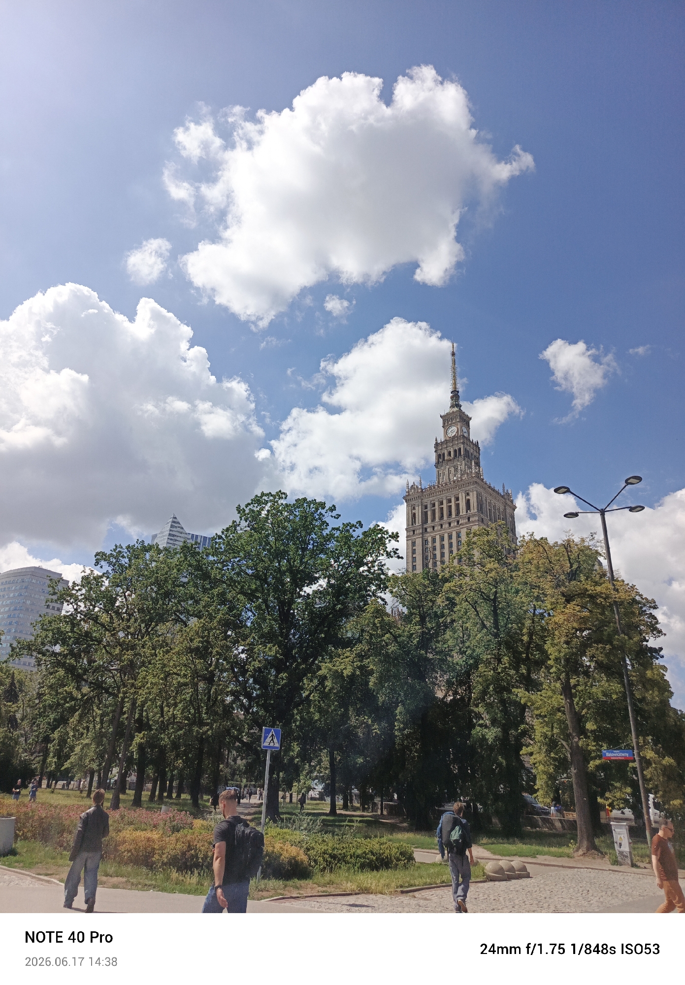This is a street-level photograph taken on a sunny day in Warsaw, Poland. The iconic Palace of Culture and Science is visible in the background, a grand, Stalinist-era skyscraper with a distinctive spire. Lush green trees with some autumn colors are in the foreground, partially obscuring the view of the building. A street lamp with multiple fixtures stands to the right of the Palace of Culture. In the lower part of the frame, a paved walkway runs through a park-like setting with grass and scattered bushes. Two individuals are walking away from the camera on the walkway. The person on the left is wearing a dark jacket and light-colored pants. The person on the right is wearing a dark t-shirt, jeans, and a backpack. A blue pedestrian crossing sign is visible further down the walkway. The sky above is a bright blue with scattered white cumulus clouds. The timestamp at the bottom of the image indicates the photograph was taken on June 17, 2026, at 14:38.