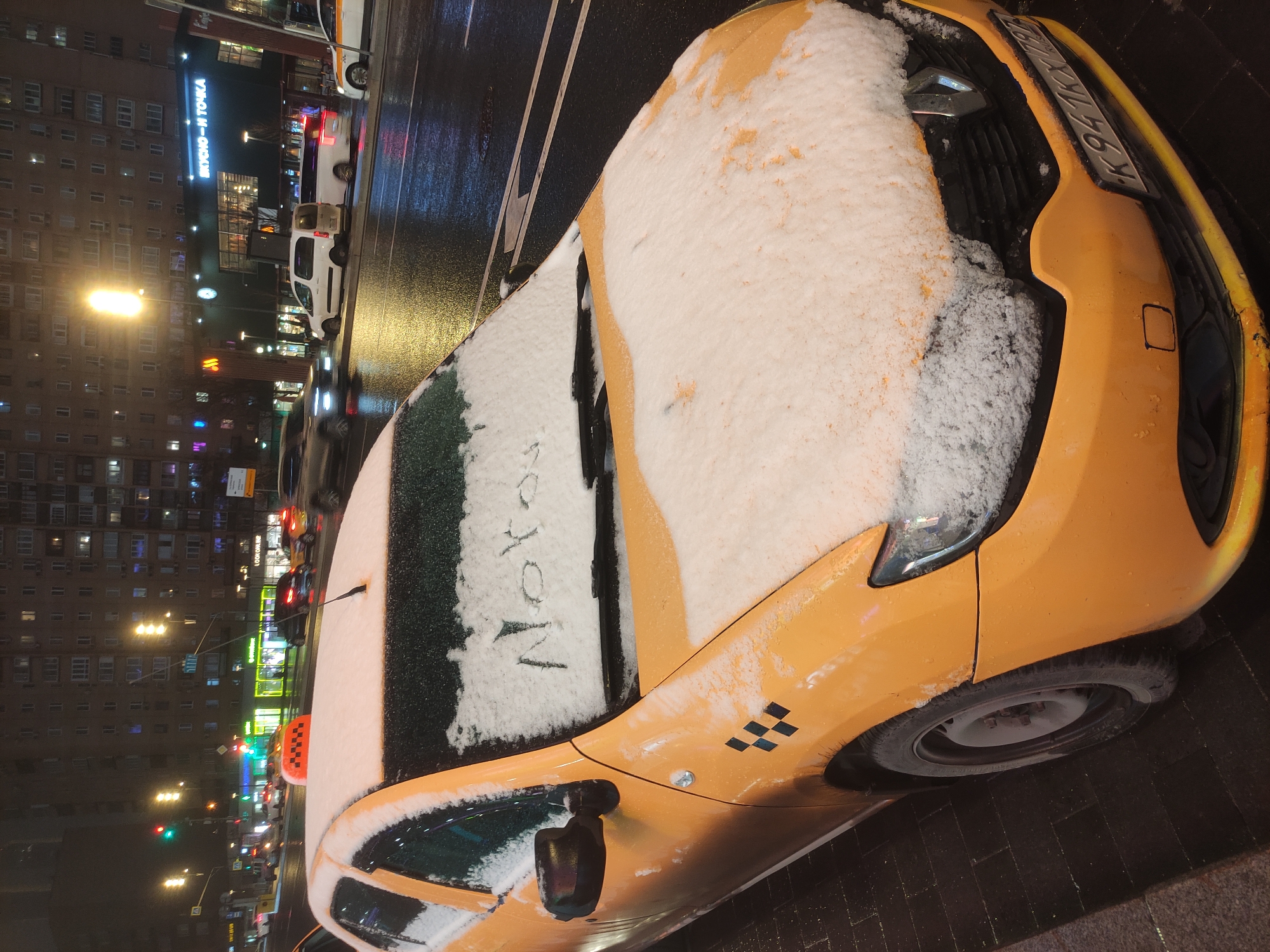 The image shows a yellow taxi parked on a wet street in Moscow, Russia, at night. The car is partially covered in snow, with the word "Moscow" written on the snow-covered windshield. The license plate reads "K941KX77". The background includes a building with a sign in Russian that reads "Вкусно - и точка" (Vkusno — i tochka), which is the Russian version of McDonald's, along with other buildings, streetlights, and traffic. The street appears to be wet and possibly icy.