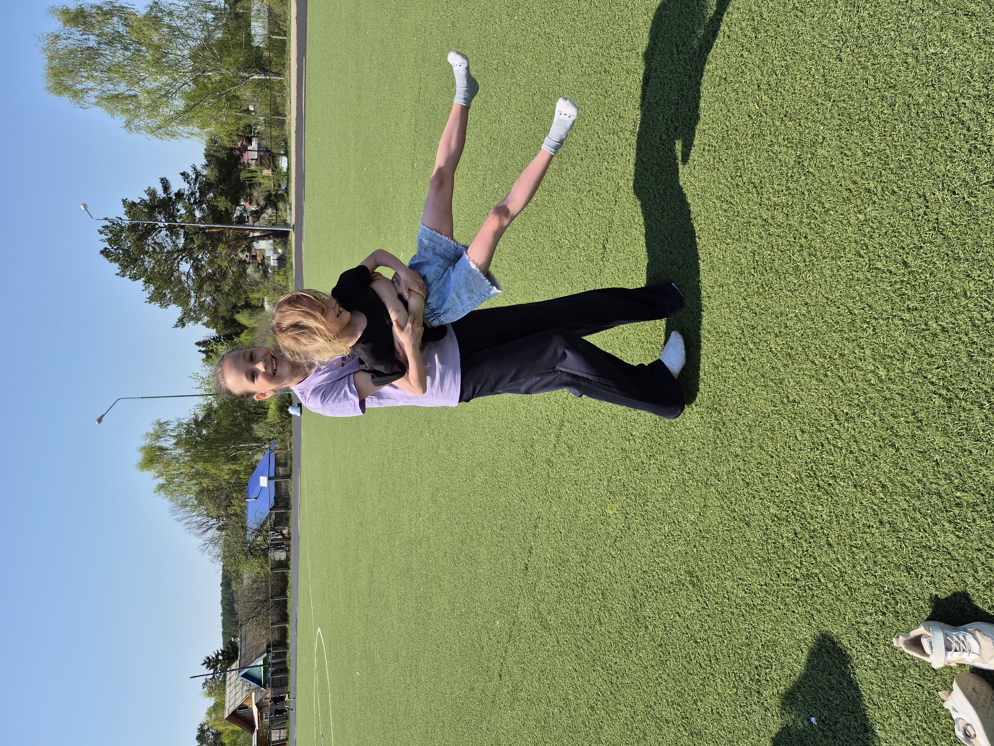 The image shows two people, likely a woman and a child, interacting on a green artificial turf field. The woman is wearing a light purple T-shirt and black pants. The child, who is held in the woman's arms, is wearing a black shirt and a denim skirt. The child's legs are extended upwards. The background includes trees, buildings, and a blue sky. Based on prior data and visual elements, the location is likely the city of Chaikovsky, Russia. Additional objects include the woman's shadow and a pair of shoes in the bottom right corner of the image.