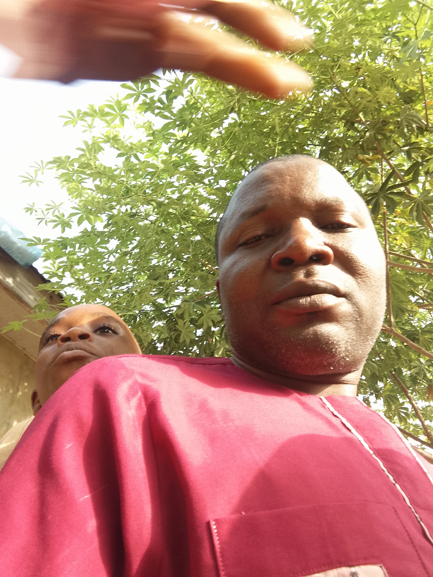 Two people are visible, though one is largely obscured. The person in the foreground is a dark-skinned man with a shaved head, wearing a dark red shirt or robe with white trim. He is looking directly at the camera. Behind him, partially obscured by his shoulder and slightly to the left, is another person with dark skin who is looking upwards and towards the left. Above the people is foliage, likely from trees. A partially visible hand is in the upper portion of the frame. The location is in the city of Unknown, Unknown.