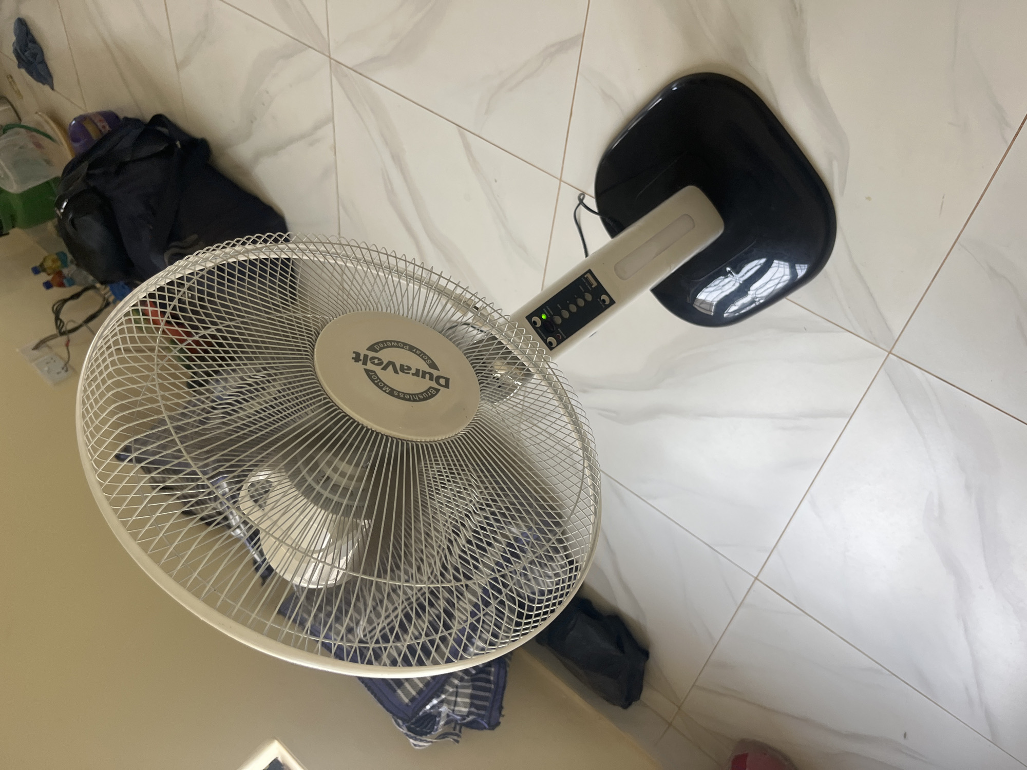 The image shows a close-up, high-angle shot of a white oscillating fan with a black base, placed on a tiled floor. The fan's grille is visible, and the center of the grille displays a circular emblem with the text "DuraVolt" and "Brushless Motor Solar Powered". The fan's stand is white, and the control panel on it is visible with several buttons and an illuminated green LED light. A black power cord is plugged into the fan and extends downwards. In the background, a dark blue backpack and other items are scattered on the floor, including some plastic bottles and a rolled-up striped fabric. The floor is covered in large, light-colored tiles with dark grout lines. The lighting appears to be from an indoor source.