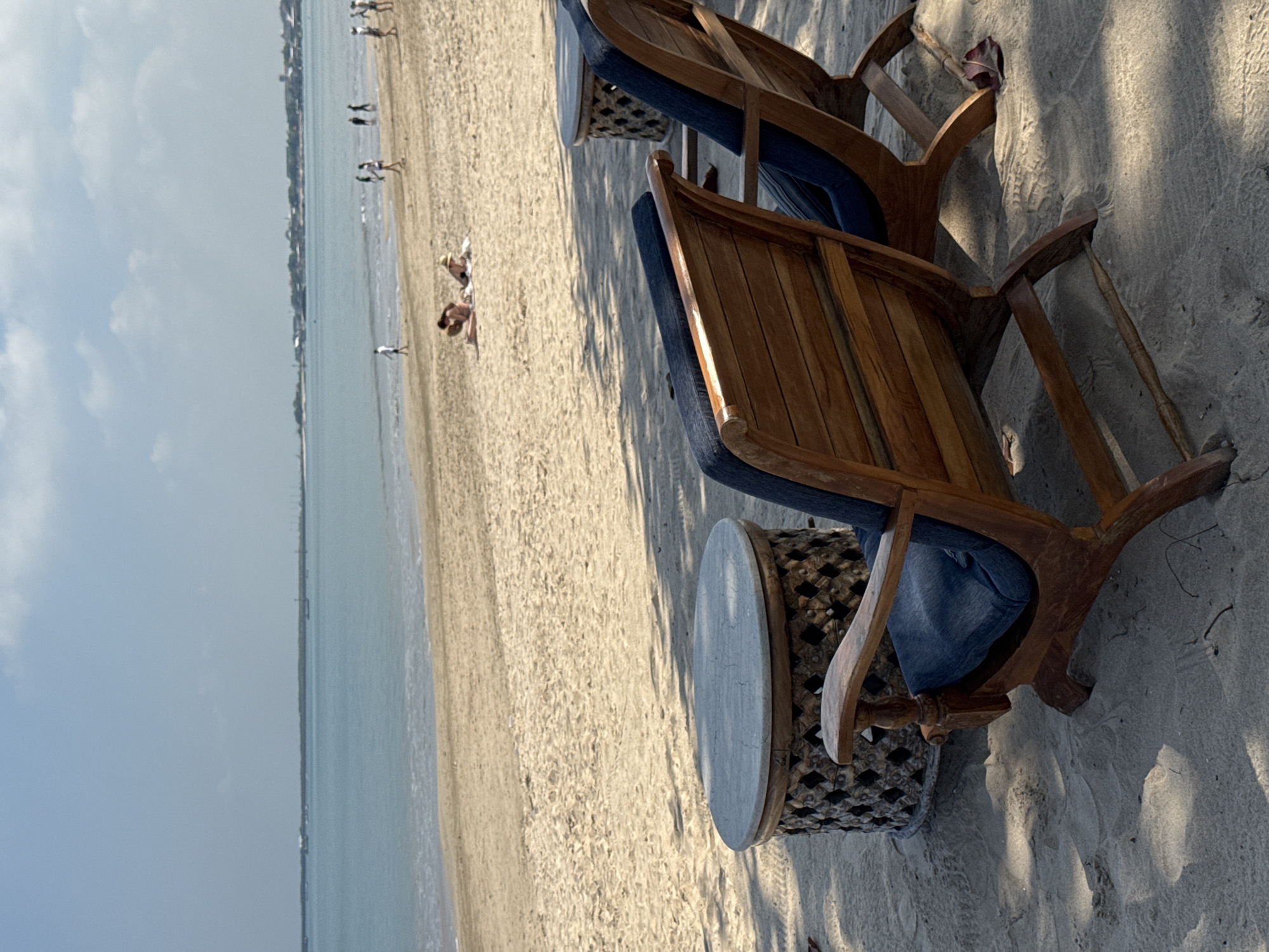 The image captures a peaceful beach scene, likely in Kuta Selatan, Indonesia, looking out towards the ocean. In the foreground, set directly on the light-colored sand, are two elegant wooden lounge chairs with blue fabric cushions. These chairs are crafted from a dark wood, featuring a slatted design on their backs and seats, with curved armrests. Between them is a small, round side table with a light-colored top and a dark, intricately patterned base.
Beyond the chairs, the sandy beach stretches a considerable distance, meeting the calm, clear turquoise waters of the ocean. Several people are scattered along the beach; some are walking near the waterline, while others appear to be lounging on the sand in the mid-ground. In the far distance, a faint coastline with subtle hints of buildings or structures is visible, suggesting a populated area or resort.
The sky is a pale blue with scattered, wispy white clouds. The lighting is bright, with subtle shadows cast by the chairs and table on the sand, indicating it is likely late morning or early afternoon. The overall impression is one of tranquility and relaxation, characteristic of a tropical beach destination. There is no visible text in the image.