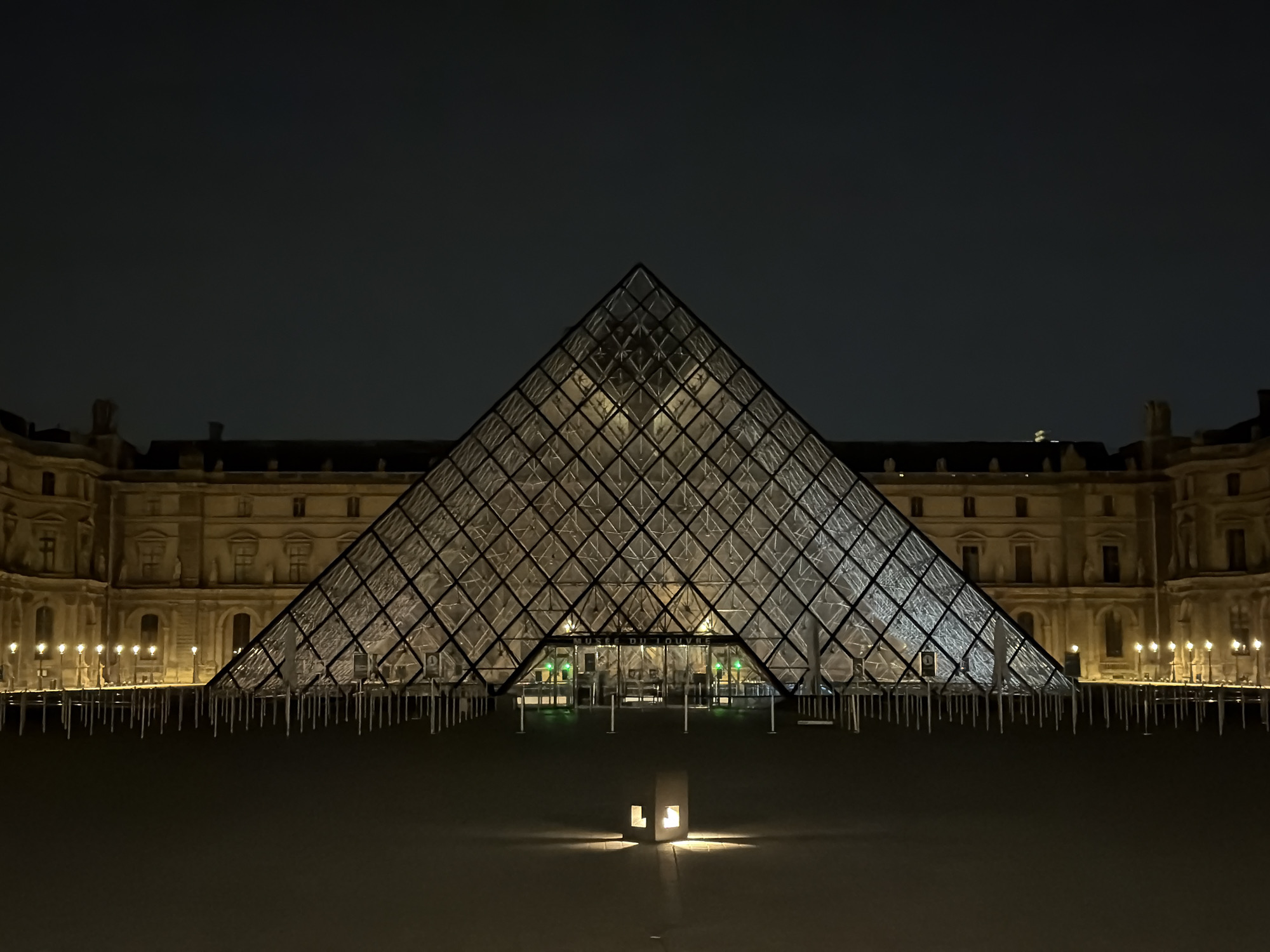 The image shows the Louvre Pyramid in Paris, France, at night. The glass and metal pyramid is illuminated from within, creating a strong contrast against the dark sky. The Louvre Museum building is visible in the background, with its facade lit by streetlights. In front of the pyramid, the courtyard is dark except for a single illuminated structure near the bottom center of the image. No people are visible.