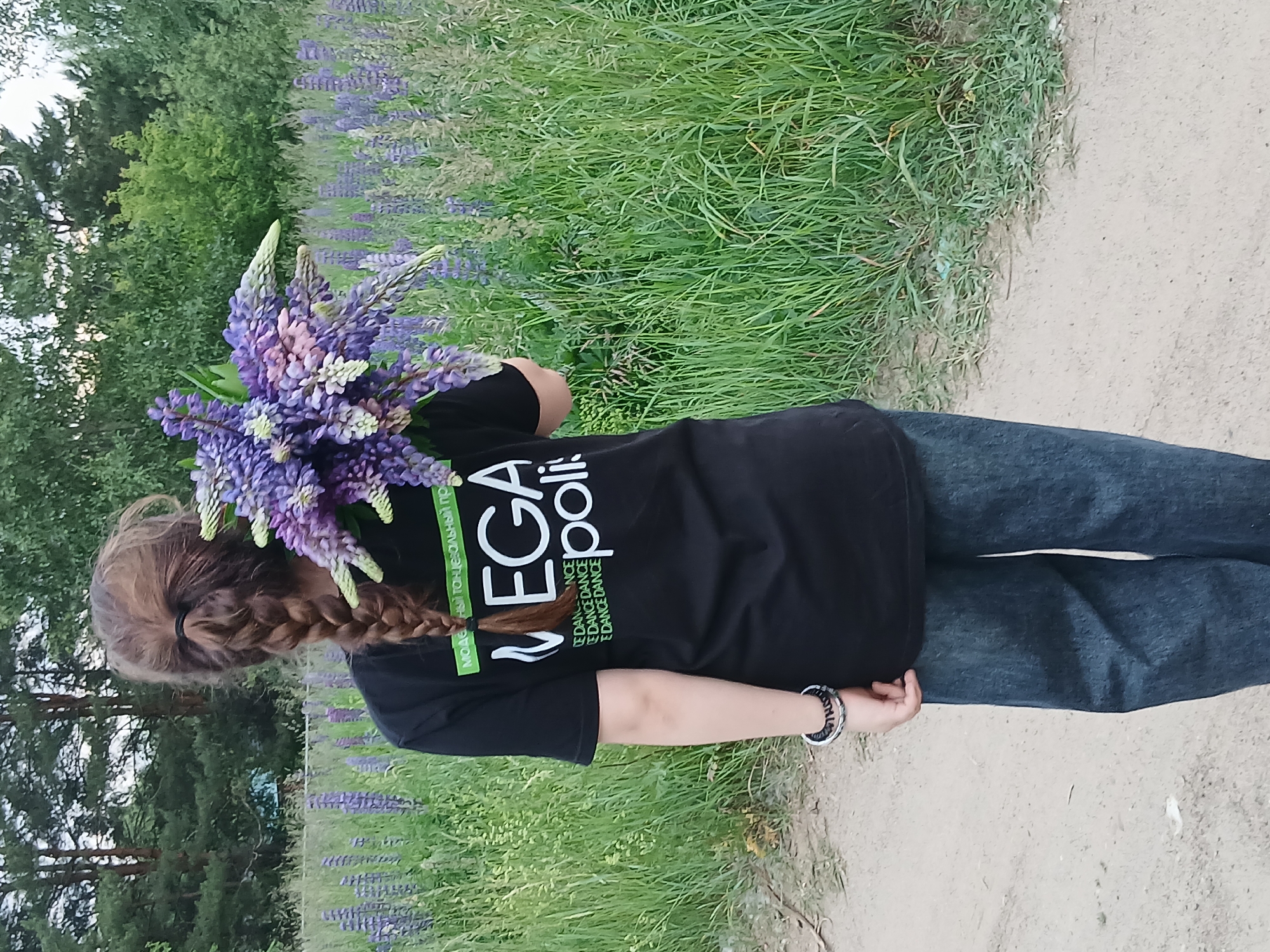 A person is standing in a field of purple lupines, holding a bouquet of lupines. The person is wearing a black t-shirt with white lettering and dark blue jeans. The t-shirt has the words "MEGA polis" on it. The person is seen from behind, with their hair in a braid. The background shows a dense field of lupines and trees. The ground in the foreground is a dirt path. The image appears to have been taken in the city of Chaikovsky, Russia, based on the lupine fields which are common in the region during the summer months.