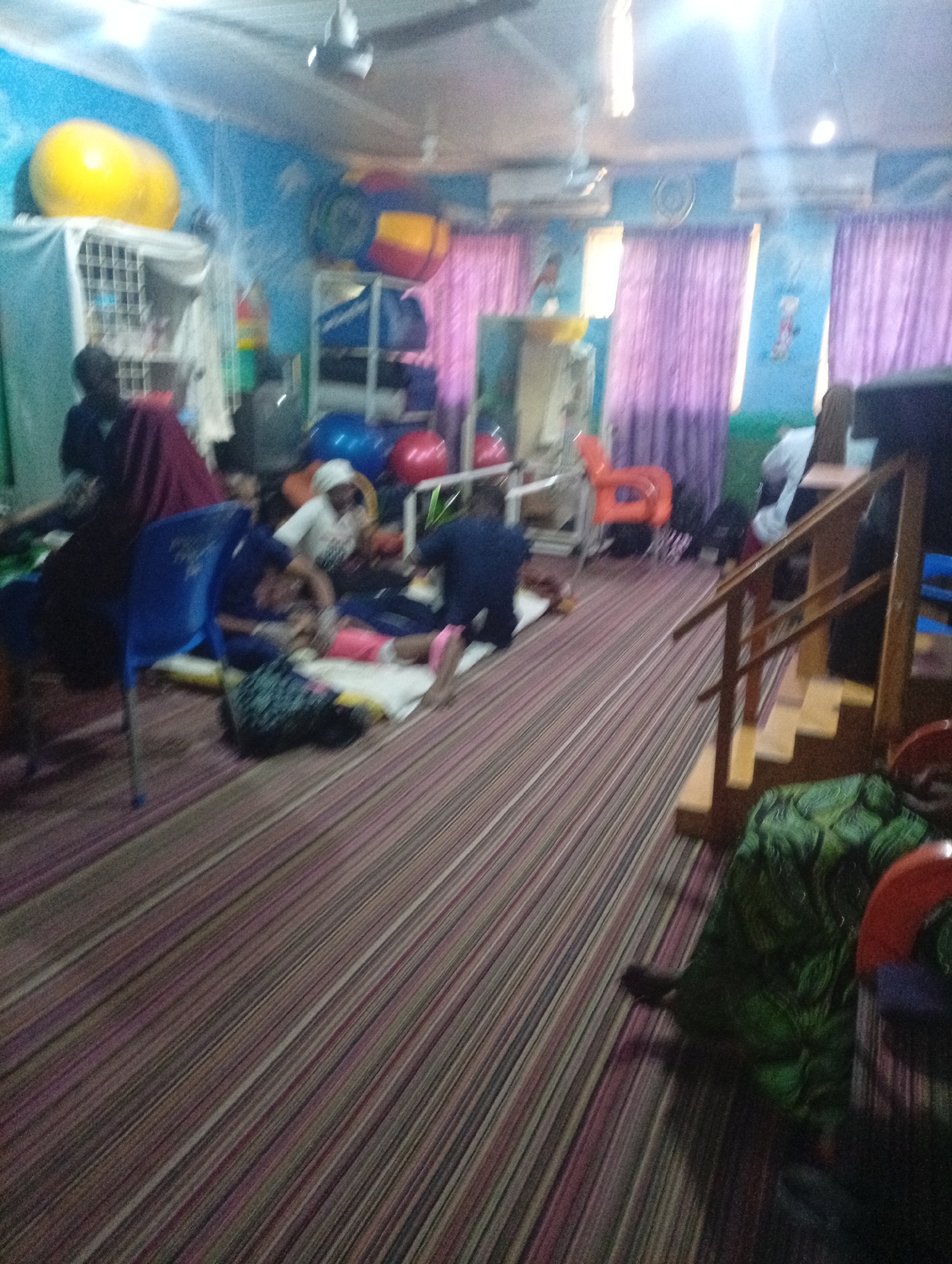 The image depicts an indoor scene, possibly a rehabilitation or therapy room, located in Gombe, Nigeria. The room is brightly colored, with blue walls and a striped carpet. There are several individuals present, some of whom appear to be patients receiving treatment. Large exercise balls in yellow and blue are visible, along with other therapy equipment like mats and chairs. Purple curtains hang across a window. The overall atmosphere suggests a functional space dedicated to physical therapy or a similar care service.
