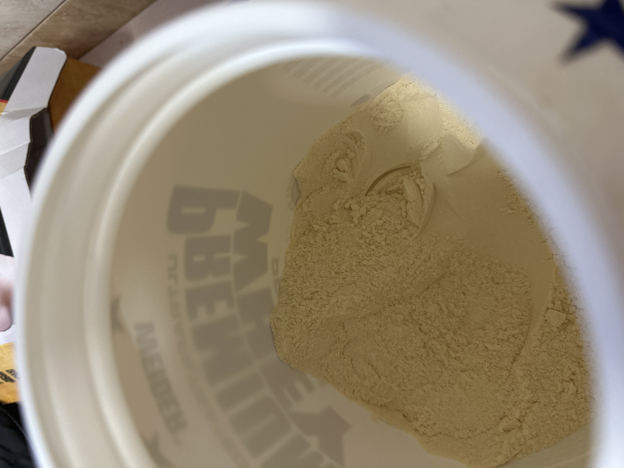 An overhead shot looks into an open white container filled with a tan powdery substance. A white scoop rests within the powder. The container has grey letters on the side. The scene takes place indoors, and a partially visible cardboard box is adjacent to the container. There is no discernible evidence to suggest the image was taken in Sofia, Bulgaria.