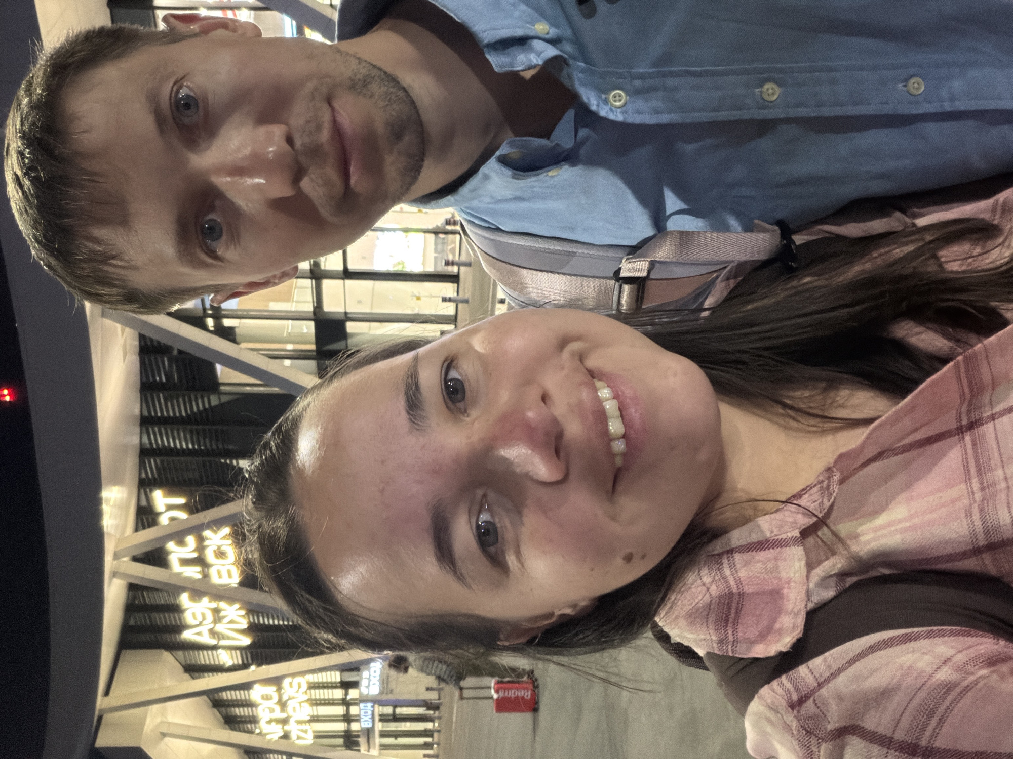 The image shows a close-up selfie of a man and a woman standing outside an airport at night. The building behind them has illuminated signage in Russian that reads "АЭРОПОРТ ИЖЕВСК" (Airport Izhevsk) and "ВХОД" (Entrance). The woman is in the foreground, smiling and wearing a pink plaid shirt. The man is to her right, looking at the camera and wearing a blue collared shirt. A red suitcase is visible in the background. The lighting suggests it is dusk or nighttime.