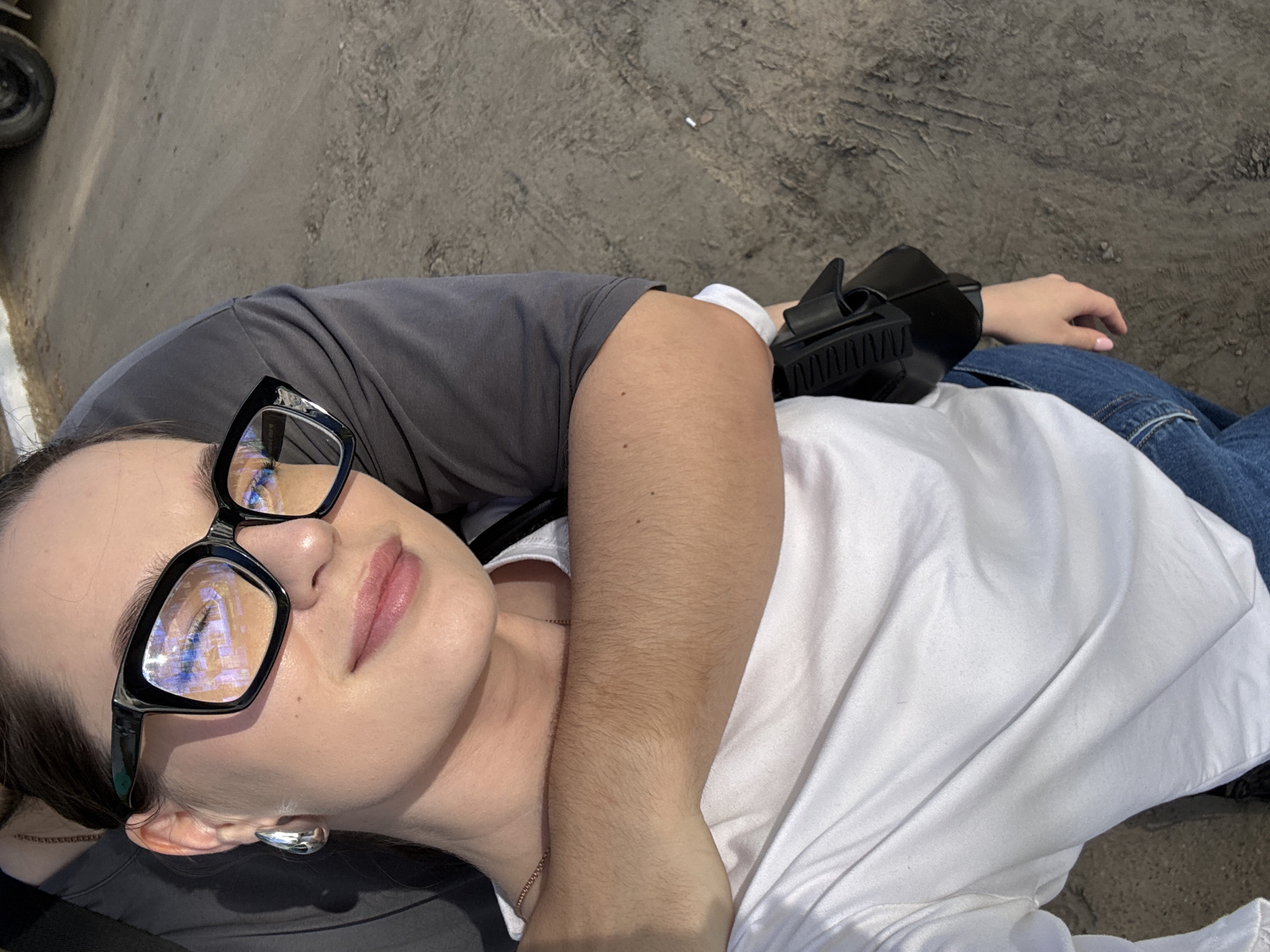 A woman is lying on the ground in what appears to be an urban or roadside setting in Chaikovsky, Russia. She is wearing large, black-framed glasses reflecting the surrounding environment. She is also wearing a white t-shirt and blue jeans. An arm, possibly belonging to another person, is draped across her. A black sandal-like object is visible near her arm. The ground is a textured, gray surface. A car tire is visible in the background.