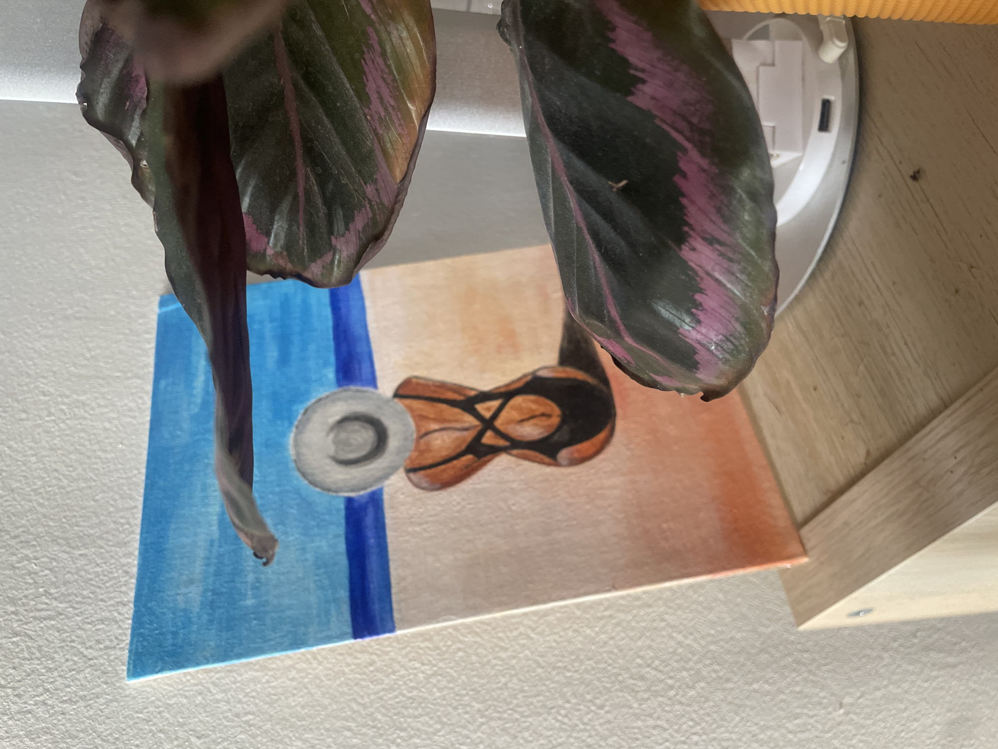 A painting of a person in a swimsuit and hat sitting on a beach is partially obscured by the leaves of a plant. The painting depicts a blue sky, a darker blue sea, and a sandy beach with an orange hue. The person in the painting is seen from behind, wearing a black swimsuit with a criss-cross back and a wide-brimmed hat. The plant in the foreground has dark green leaves with bright pink markings. The painting is placed on a light-colored wooden surface, possibly a shelf or table. The background is a textured white wall. There are no visible signs or indicators that would suggest a specific location in Atarfe, Spain.