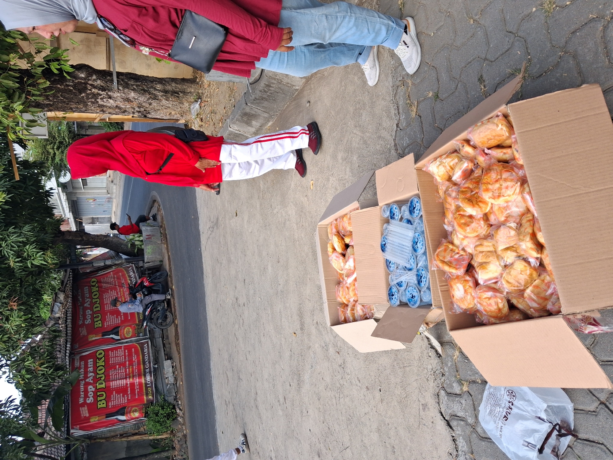 In Surakarta, Indonesia, two women are standing on a sidewalk next to two cardboard boxes filled with food. The woman on the left is wearing a red hijab and a red shirt with white pants. She has a black purse slung across her body. The woman on the right is wearing a gray hijab and a maroon shirt with light blue jeans. She also has a black purse.
The cardboard boxes are full of individually wrapped food items, which appear to be pastries or bread. One box also contains several small bottles of water and plastic straws. In the background, a banner hangs on a building advertising "Warung Makan Sop Ayam BU DJOKO". A motorcycle is parked in front of the banner, and a man is sitting on it. The sidewalk is made of cobblestones and there is a street running alongside it. Trees and foliage are visible in the background, suggesting a semi-urban or suburban environment.
