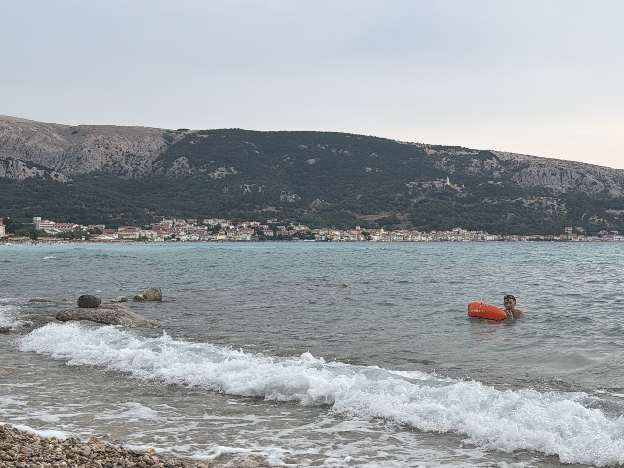 The image captures a coastal scene in Općina Baška, Croatia, under an overcast sky. In the foreground, a wave is breaking on a pebble beach, with a section of the beach visible at the bottom left. The sea stretches out into the middle ground, where a person is swimming while holding onto an orange inflatable buoy labeled "NABAIJI". The person appears to be looking towards the camera. In the background, a town with numerous buildings is nestled along the coast, backed by a large, tree-covered mountain. The mountain range extends across the horizon. The water is a muted blue-green.