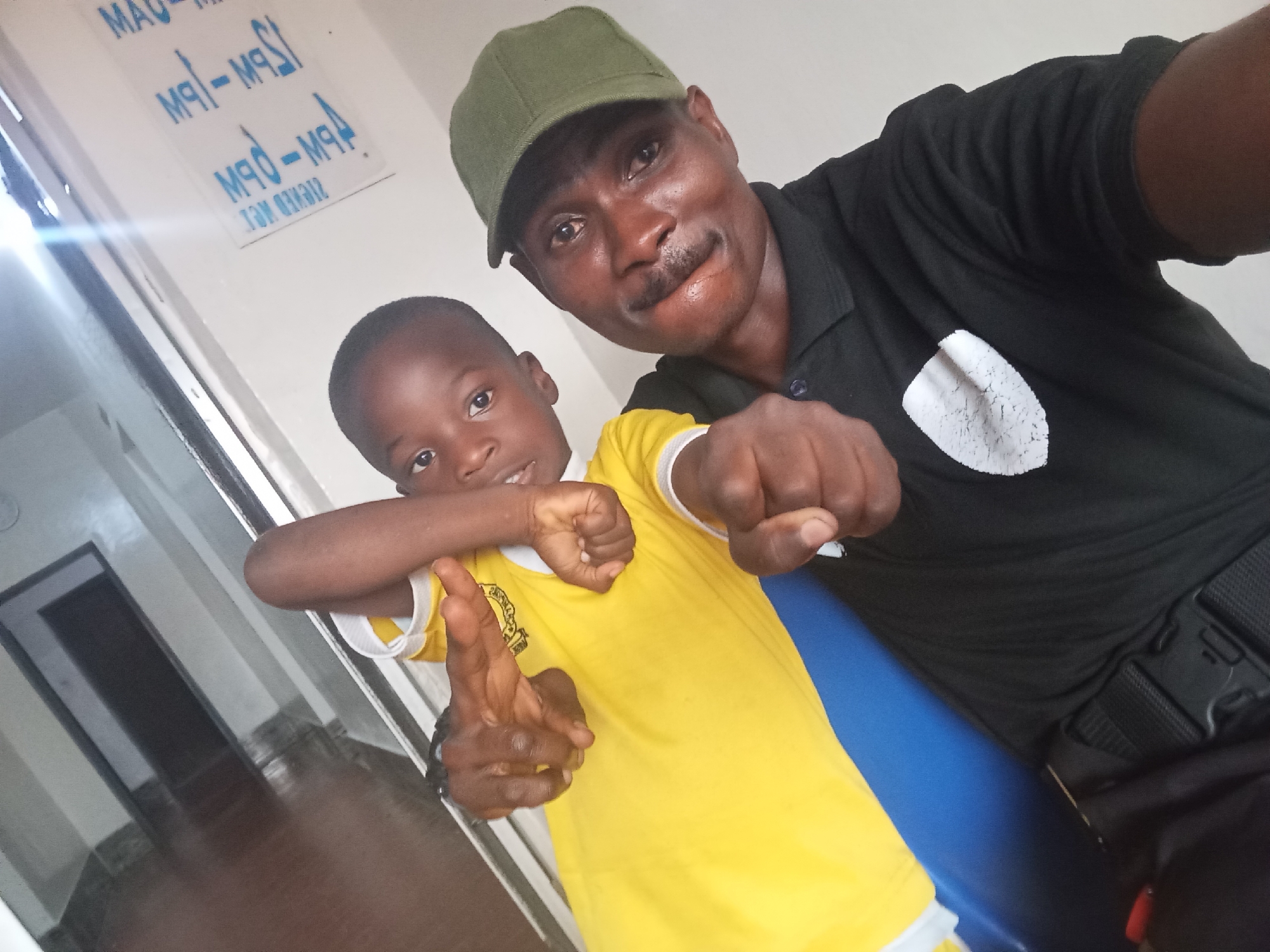 In this photograph taken in Jos, Nigeria, two individuals, an adult male and a young boy, pose for a selfie. The adult male, wearing a green cap and a black polo shirt with a white emblem, stands on the right, his right arm extended towards the camera in a fist. The young boy, dressed in a bright yellow t-shirt with a white collar and a crest on the left chest, is positioned to the left of the adult. He has his left arm bent, with his fist raised, and his right hand is extended, palm open, in a gesture resembling finger guns. Both subjects are looking towards the camera. The background features a white wall with blue signage displaying text in what appears to be a reversed, possibly mirrored, format, indicating a reflective surface or a printing error. The lighting suggests an indoor setting, possibly a hallway or a room with a door visible on the left. The overall scene captures a casual and playful moment between the two individuals.