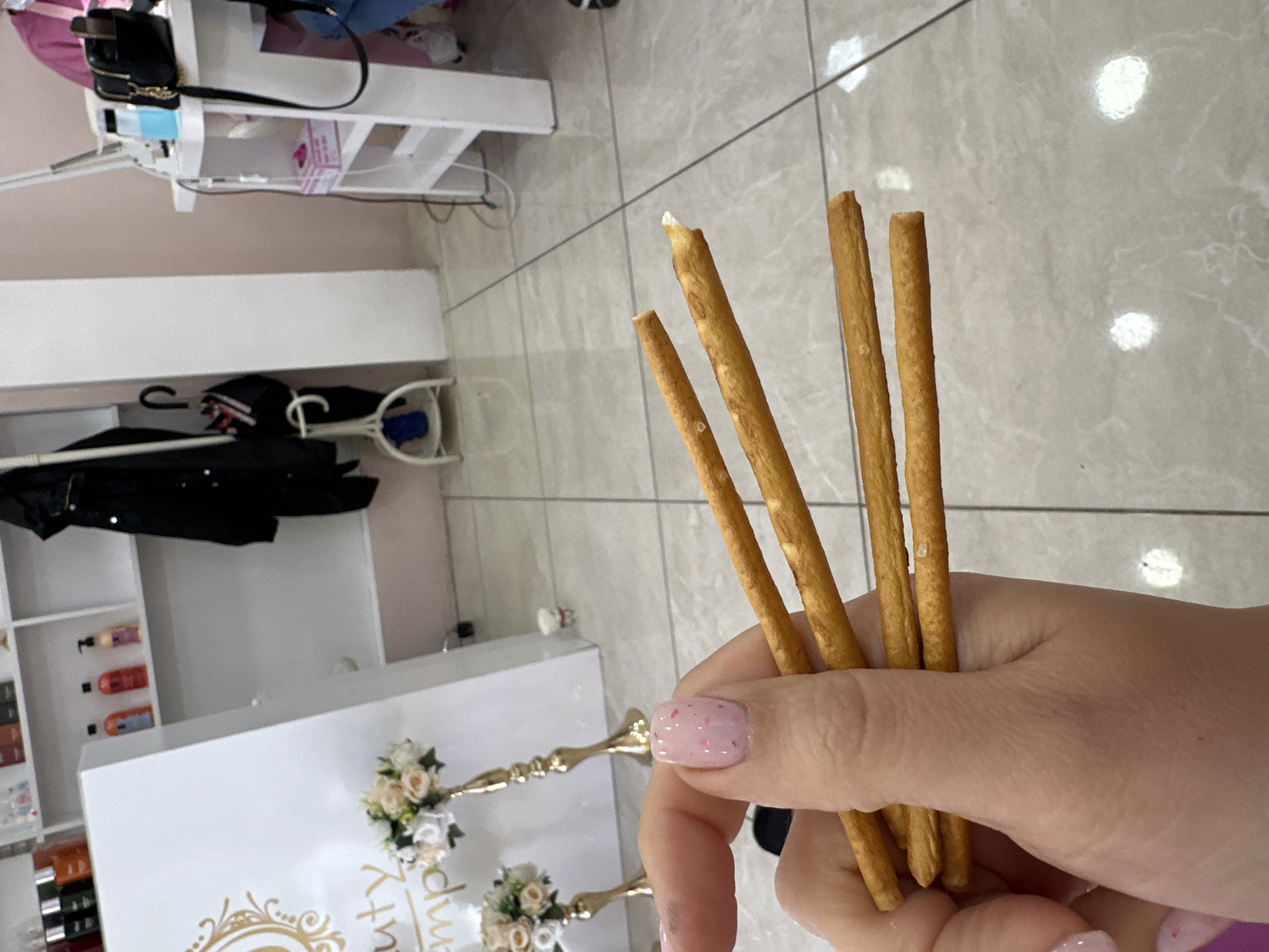 The image displays a close-up of a person's hand holding five golden-brown breadsticks, likely grissini or pretzel sticks, in an indoor setting. The hand is adorned with pink nail polish featuring small, lighter pink specks. One breadstick appears broken at the top, revealing a lighter interior, and some salt crystals are visible on their surface.
The background reveals the interior of what strongly appears to be a beauty salon or similar commercial establishment. On the left, white shelving units are visible, holding various bottles and beauty products. A black handbag rests on a shelf. Below the shelves, a coat rack with a black jacket can be seen. In the lower-left quadrant, a white table or counter features a small floral arrangement of cream and white roses, held in golden candle holders or decorative stands. Partially visible golden text on the table reads "Beauty Emp...", suggesting a name like "Beauty Empire" or "Beauty Emporium," which further supports the salon environment. The flooring is light-colored tile with visible grout lines. The lighting suggests it is daytime. The scene captures a moment of waiting or relaxation, perhaps while enjoying a snack in a professional service setting in Sofia, Bulgaria.