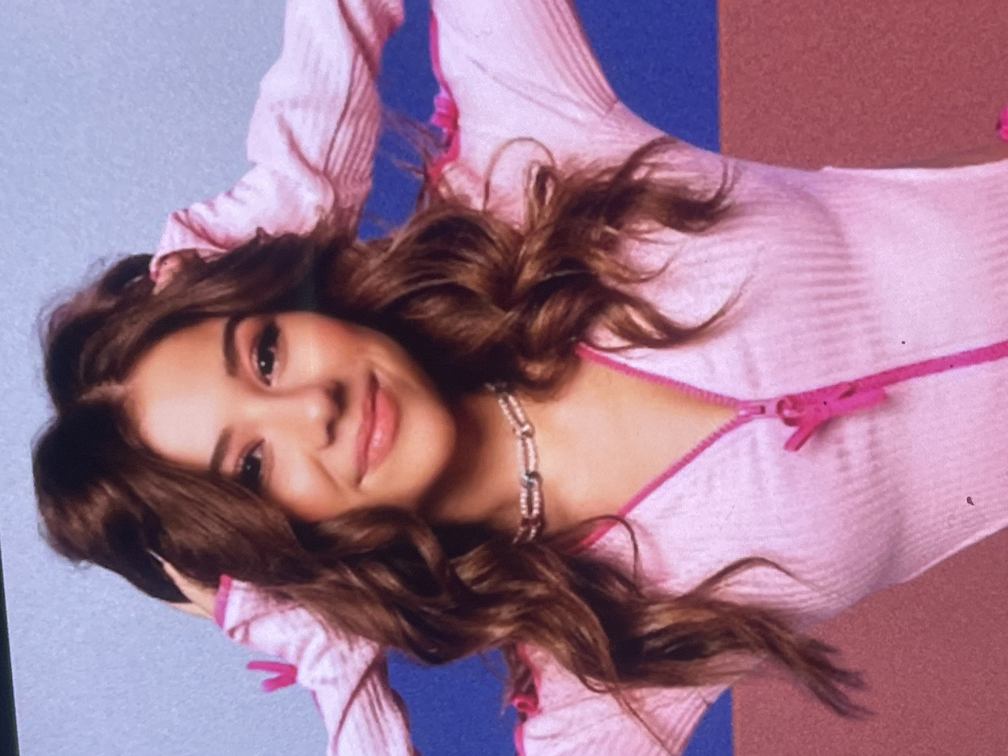 This media file, situated in Poltava, Ukraine, presents a portrait of a young woman. The image is rotated 90 degrees counter-clockwise, showing the woman's head on the left and her body extending to the right. She has long, voluminous, wavy dark brown hair that cascades around her shoulders and back. She is looking directly at the viewer with a gentle, relaxed smile.
The woman is wearing a light pink, possibly ribbed, long-sleeved top or dress. A prominent, bright pink zipper runs down the front of the garment, featuring a matching pink zipper pull. Around her neck, she sports a distinctive, chunky chain-link necklace, which appears metallic and may have small, reflective accents. Her makeup is natural, enhancing her features.
Her arms are positioned behind or above her head, creating a relaxed pose. The background is a studio-style setup, featuring distinct blocks of color: a soft light blue section is visible to the upper left, transitioning into a royal blue area, and then into a dusty rose or reddish-brown section on the lower right. The overall impression is a vibrant and styled portrait. There is no visible text in the image.