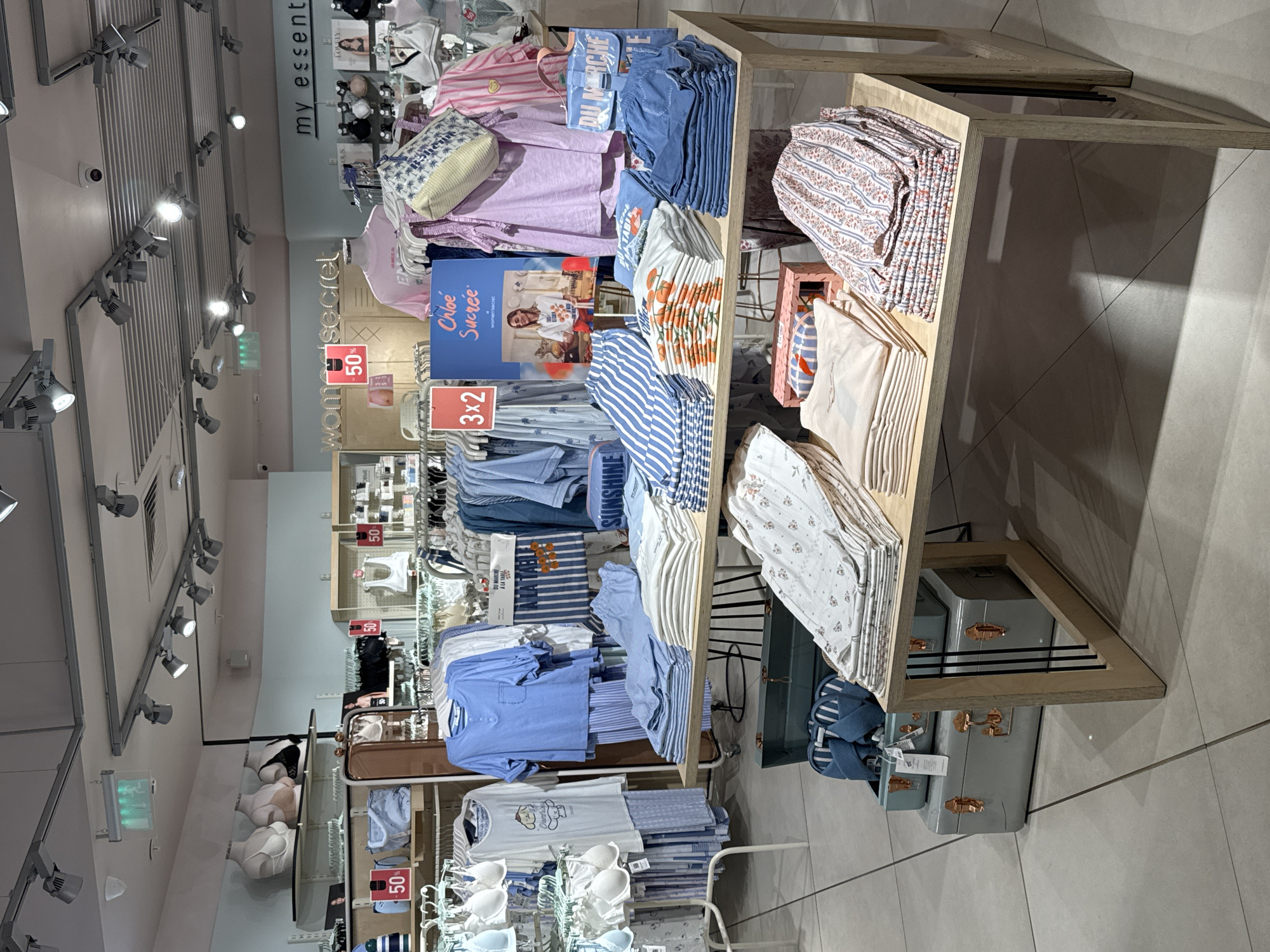 A clothing display in a retail store.
**Environment:**
The image shows the interior of a well-lit retail space, likely a clothing store. The lighting is primarily from overhead track lighting, common in such environments.
**Objects:**
The main feature is a multi-tiered display table filled with folded clothing, including tops, pants, and lingerie, arranged by color and style. Price and promotional signage are attached to some of the clothing racks.
**Interaction:**
The items are arranged in a manner to attract customers, indicating commercial activity.