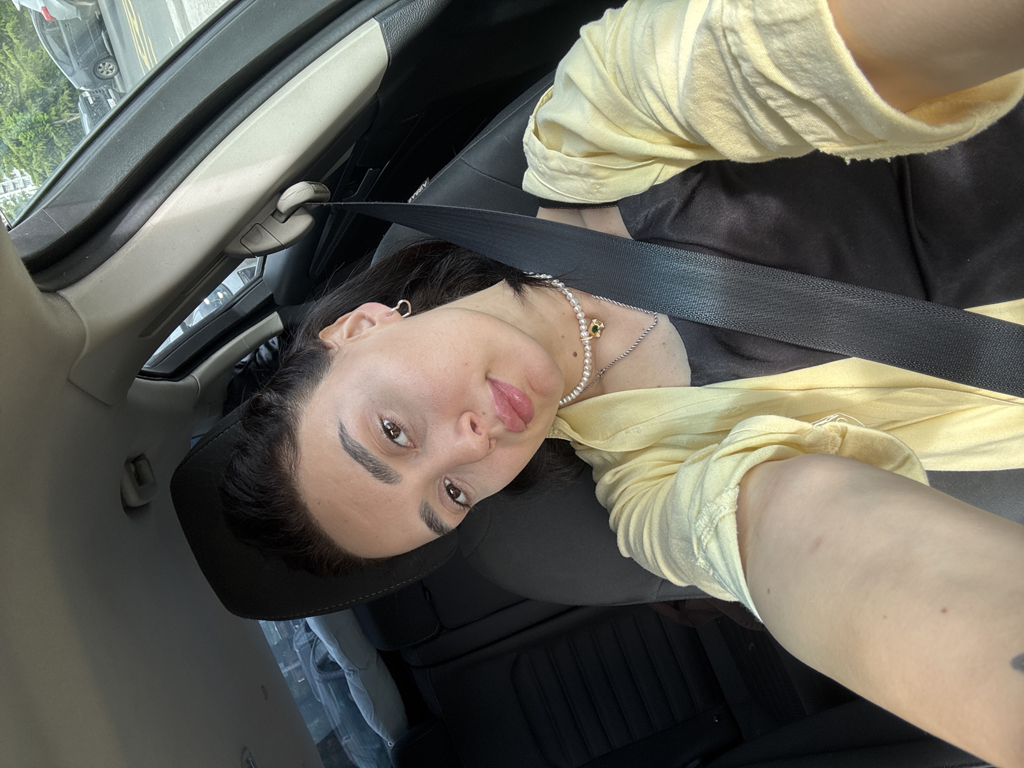 A young woman is seated in the driver's seat of a car, wearing a black shirt and light yellow jacket or shirt, fastened with a seatbelt. She's wearing a necklace of pearls and silver chain. She has dark hair and eyebrows, fair skin, and light brown eyes. The car's interior is dark, with black leather seats and a beige roof. A sunroof is visible, showing a street scene outside with parked cars and greenery. The image is taken at an unusual angle, perhaps by the woman herself. The location is plausibly Tbilisi, Georgia, judging from the combination of vehicle and cityscape visible through the sunroof.
