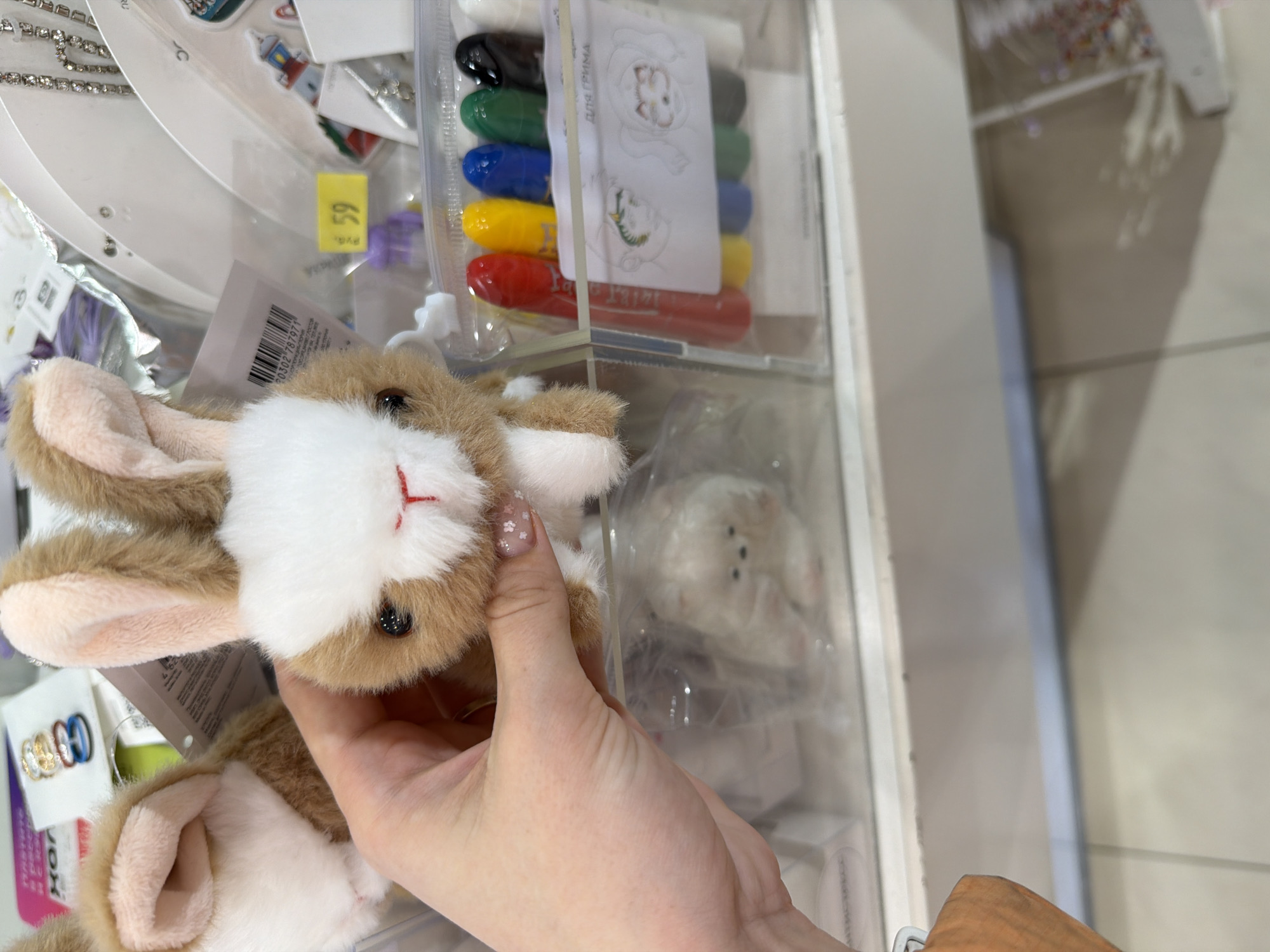 A person is holding a small, plush rabbit toy in their hand within what appears to be a retail setting. The hand has a visible manicure. The rabbit is brown and white with a red embroidered nose. Behind the rabbit, there are various packaged items on display in a clear shelving unit. These items include what appear to be colored crayons or markers, and possibly other toys or accessories in plastic packaging. The retail setting is indoors, with the shelf constructed of a white material. Based on the information in the prompt, this retail setting is located in the city of Berezniki, Russia.