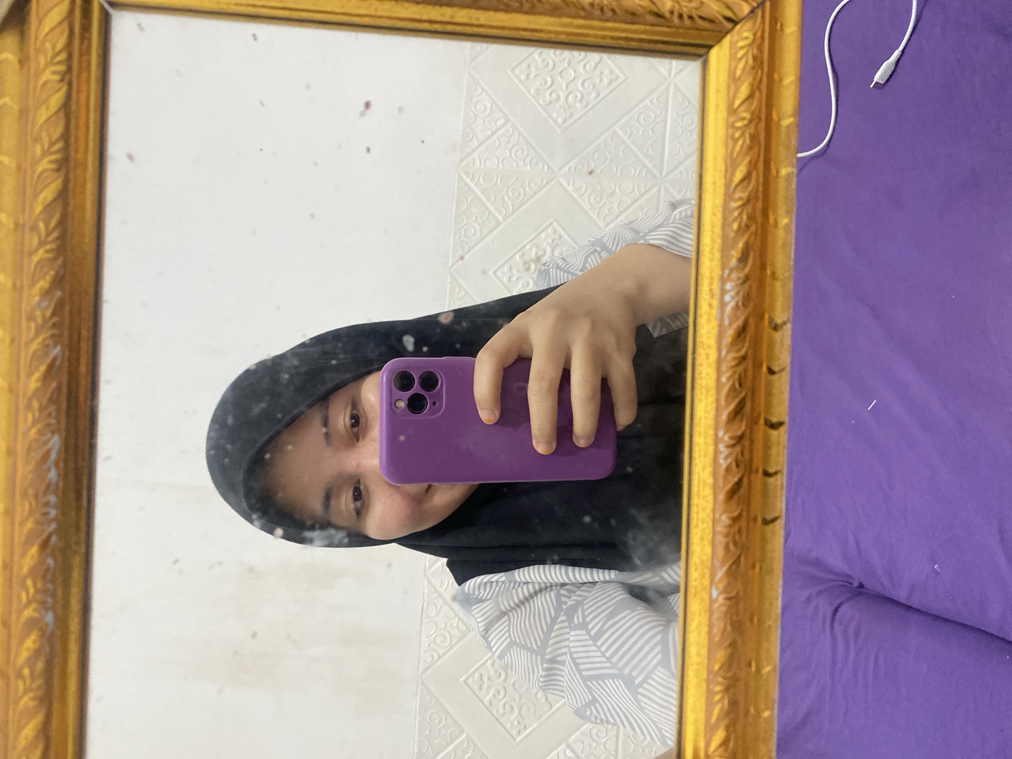 A young woman, wearing a black hijab and a grey and white patterned shirt, takes a selfie in a gilded mirror. She holds a purple smartphone in her right hand, which is positioned in front of her face. The mirror reflects a white wall with a raised, geometric pattern. The foreground shows a purple surface, possibly a cushion or bed, with a white charging cable resting on it. The scene appears to be indoors, likely a bedroom, in Banjarbaru, Indonesia.