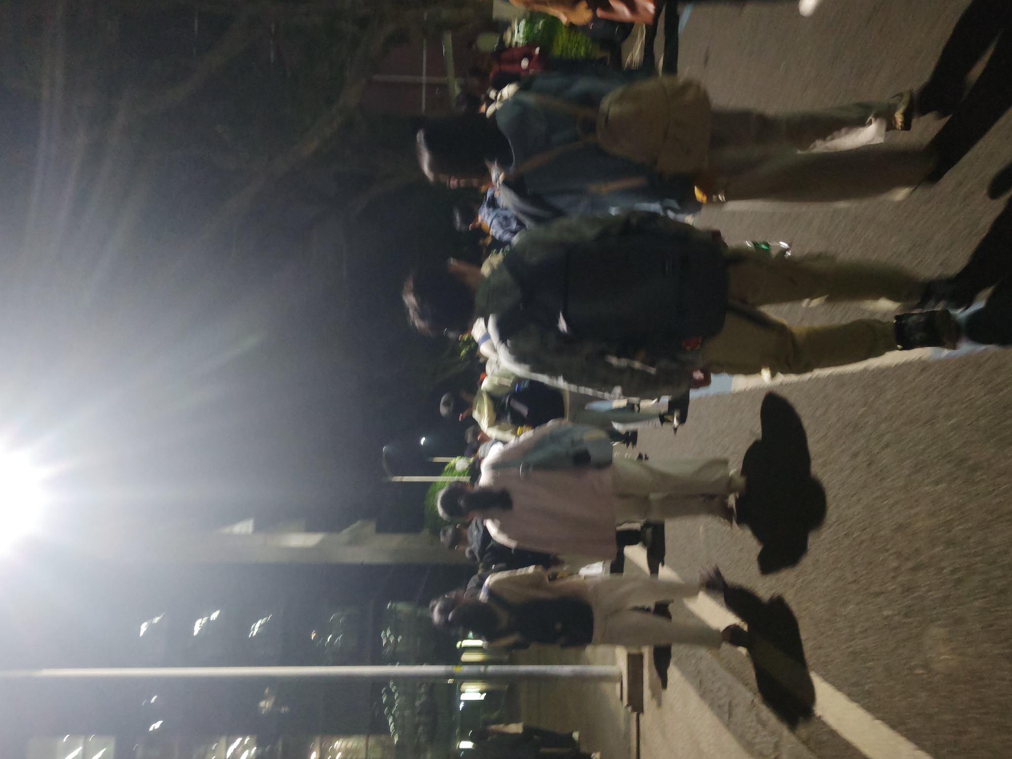 A nighttime street scene in Chongqing High-tech Zone, China shows a group of people walking on a sidewalk. They are likely students walking at night as their backs are to the camera. Some are wearing backpacks and casual clothing. The scene is illuminated by streetlights. The building is a dormitory for Chongqing University. Shadows of the people are cast on the asphalt sidewalk.