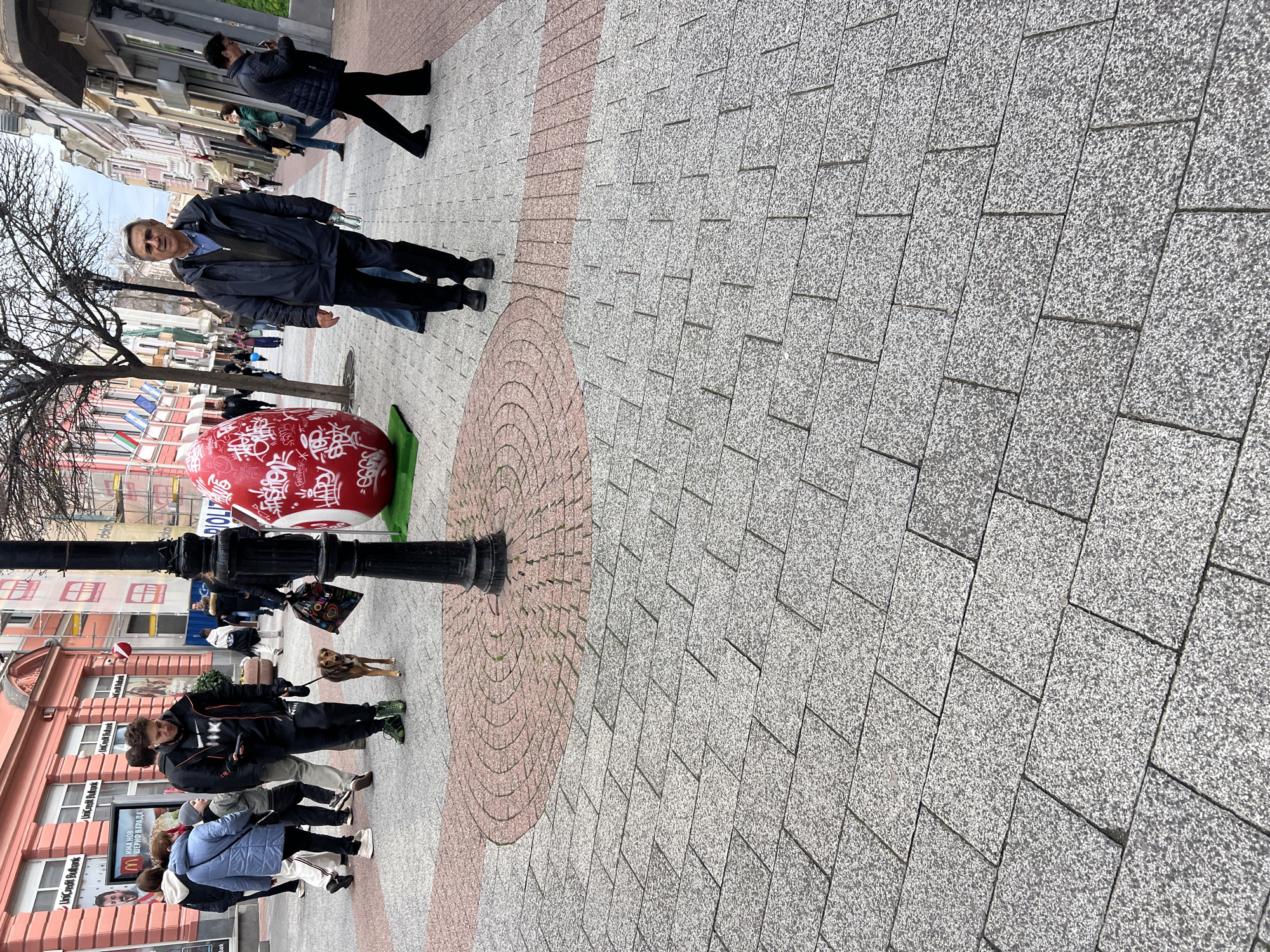 The image captures a street scene in Plovdiv, Bulgaria. The foreground features a paved walkway with a distinctive pattern of gray and pinkish-red interlocking stones. A large, red, egg-shaped sculpture adorned with white calligraphy stands near a black lamppost. A man in a dark blue jacket and black trousers is standing next to the egg. Further down the street, a group of people are clustered, including a person walking a dog on a leash. Buildings line the street, featuring businesses with signs in Bulgarian. Additional pedestrians can be seen in the distance. The overall atmosphere is that of a typical urban street scene with a public art installation.