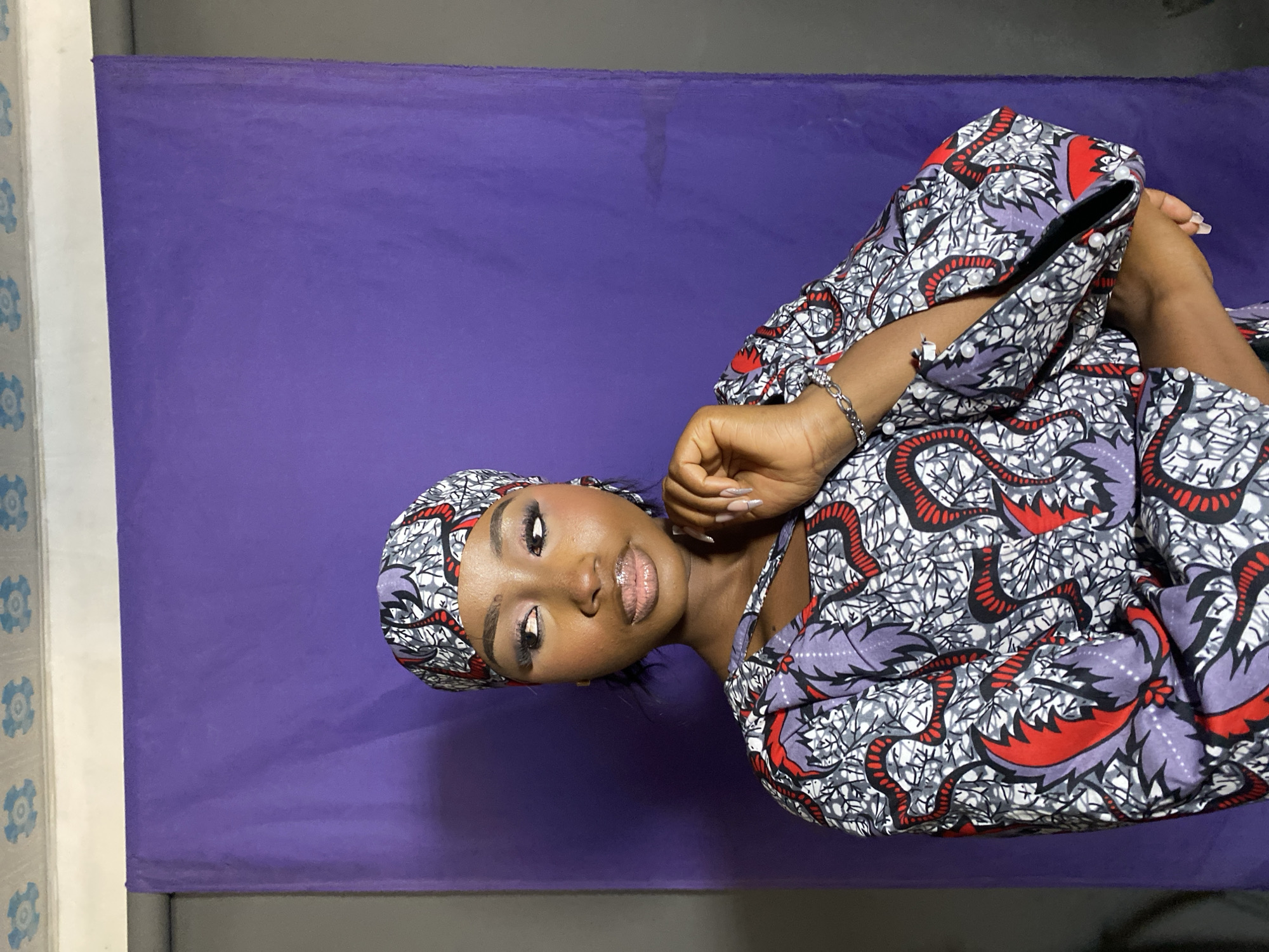 A woman is facing the camera, wearing a patterned top and headwrap, posing against a purple backdrop. The location is in the city of Gura Top, Nigeria.