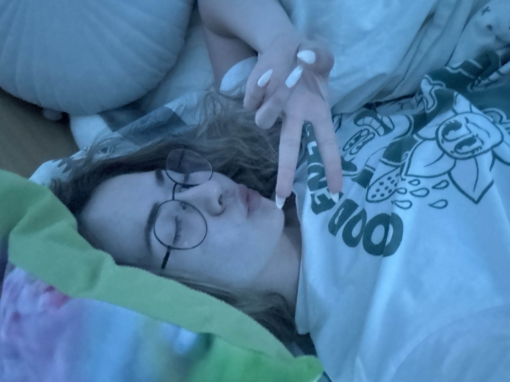 A young person with glasses is lying in bed. The person is facing the camera, with their eyes closed. Their left hand is raised, forming a peace sign. They appear to be wearing a t-shirt with cartoonish designs. There is a pillow behind them, and colorful bedding partially visible. The image seems to be taken in a domestic setting, most likely a bedroom. There is no evidence in the photo to indicate this is in Blagoevgrad, Bulgaria, or any other specific location.