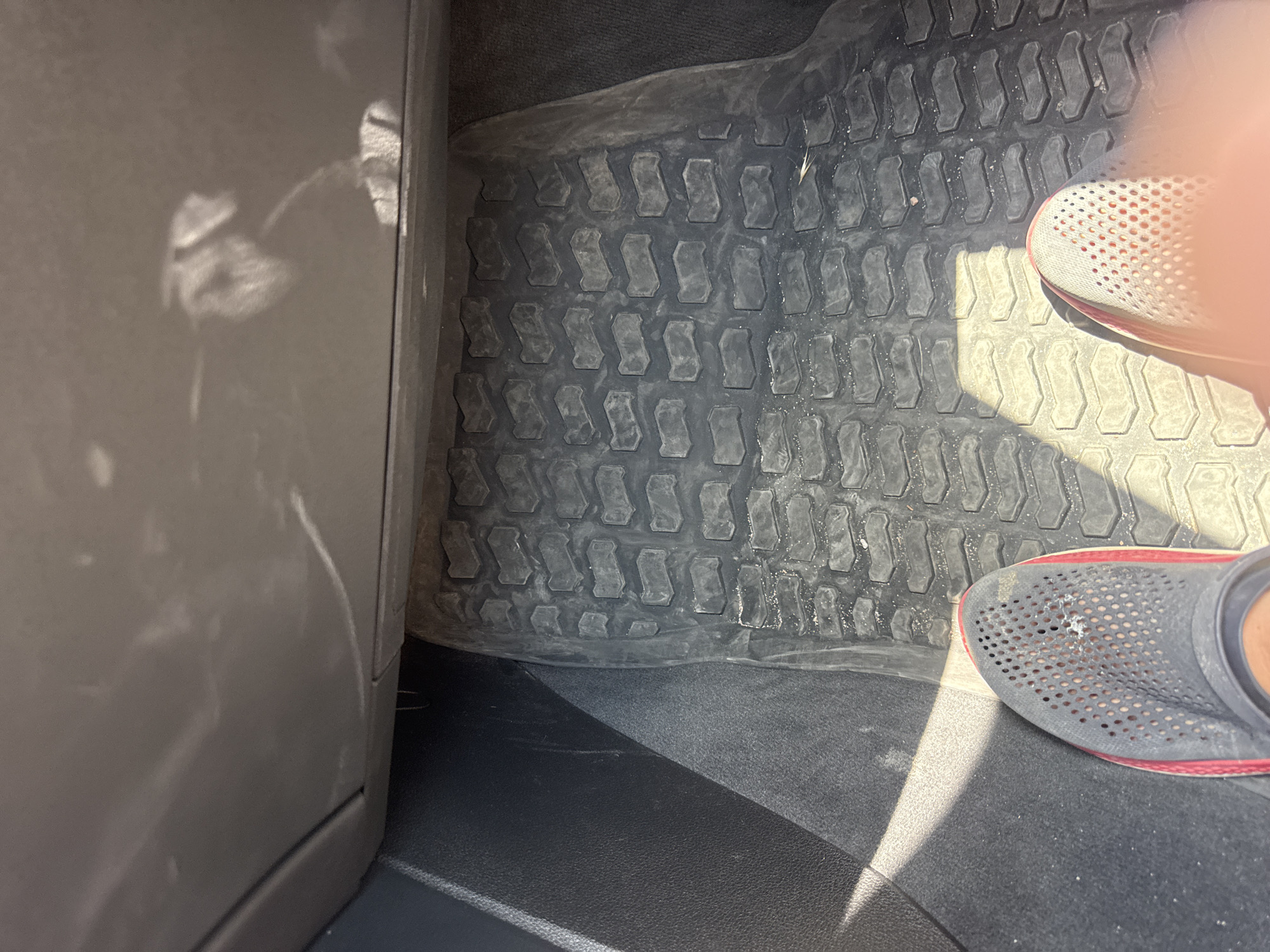 The image shows a close-up view of a vehicle's floor mat, likely inside a car. The mat is black with a textured, tire-tread-like pattern. Sunlight casts distinct shadows across the mat, indicating it's daytime, likely a sunny day. On the right side of the frame, a person's feet are visible, wearing dark gray clogs with red trim and a perforated upper. One shoe is partially obscured by the leg of the person. The shoes are resting on the floor mat. To the left, a portion of the car's interior trim is visible, appearing to be a dark gray or black plastic panel. The overall scene suggests a casual, everyday moment, possibly while parked. The location is identified as Boreti, Montenegro. No text is visible in the image.