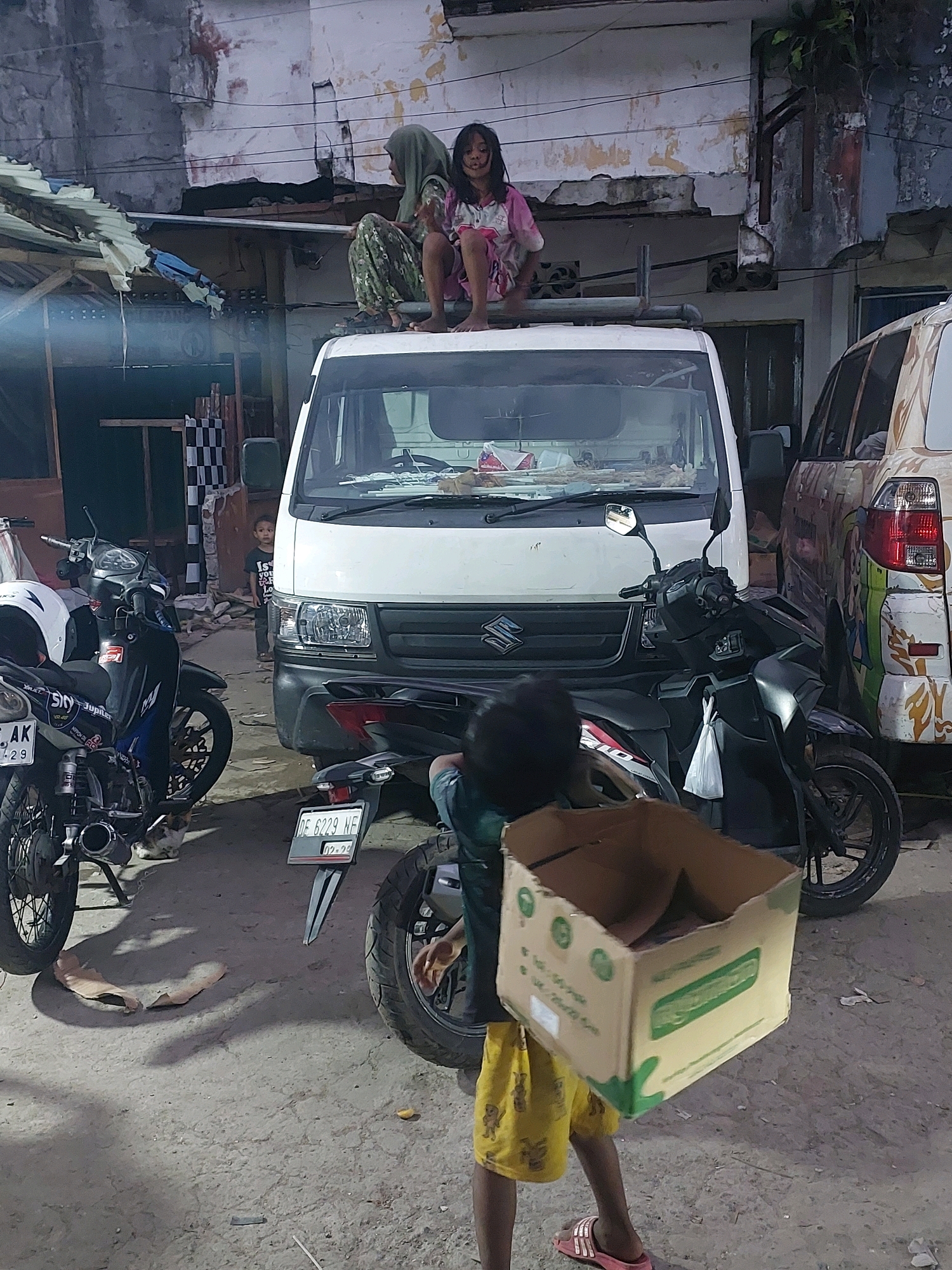 Two young girls, one wearing a hijab and the other a pink dress, are sitting on top of a white truck in Ambon, Indonesia. A young child, wearing a dark t-shirt and shorts, stands near the front of the truck. Two motorcycles are parked on either side of the truck. A cardboard box is being carried by someone out of frame in the foreground. The background appears to be an urban environment with dilapidated buildings and debris. The lighting suggests it is either dawn, dusk, or an overcast day.