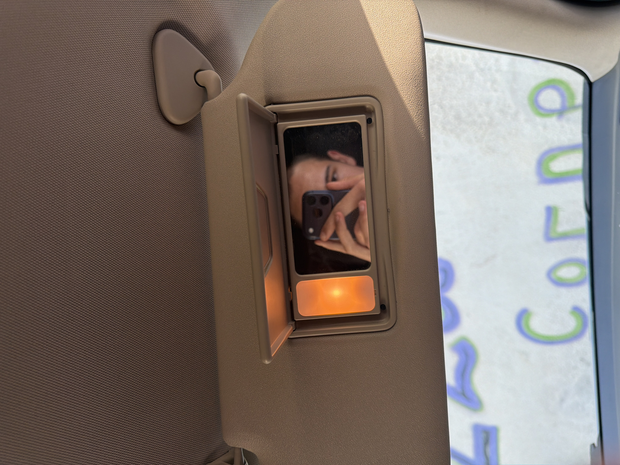 A close-up, slightly high-angle shot captures the interior of a car's visor, which is flipped down. The visor is a light beige color and has a textured surface. Within the visor, there is a mirror that reflects a person's face as they hold up a phone to take a selfie. To the left of the mirror is an illuminated light, emitting a warm orange glow. The background outside the car's windshield is blurred, showing what appears to be writing on a surface. The writing consists of large, colorful letters in blue and green, with partial words visible. The overall impression is that of a casual self-portrait taken inside a vehicle.