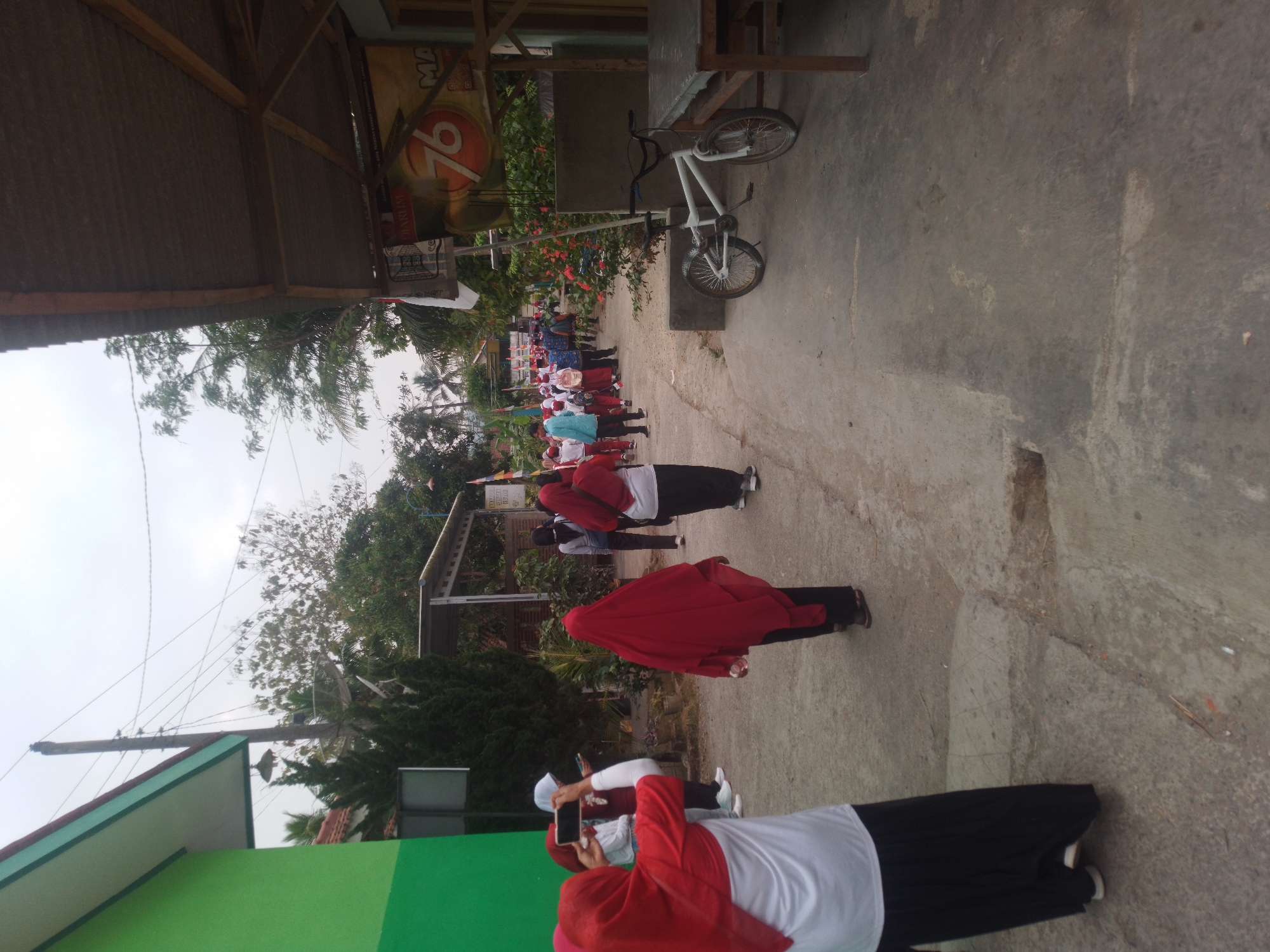 The image depicts an outdoor scene, likely a street or pathway, in the city of Kebagoran, Indonesia. In the foreground, two individuals are visible. One person on the left, wearing a red and white hijab and a black skirt, is holding a smartphone and appears to be taking a picture. Another person, also wearing a red hijab and a black skirt, is walking away from the camera.
Further down the pathway, a group of people, predominantly women and girls wearing hijabs and uniforms, are walking in a procession. Some are carrying flags. The environment includes buildings, trees, and a bicycle parked next to a table. A large banner with the number "76" and the word "DIARUM" is visible on the right side of the image. The sky is overcast.