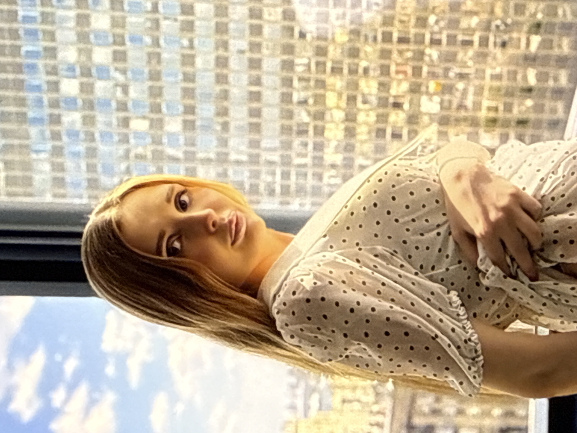 The media file shows a young woman lying down, looking directly at the camera. She has blonde hair and is wearing a white polka dot dress with ruffled sleeves. Her hand is resting on her dress. The background is out of focus, but appears to be an urban setting with a tiled wall and a sky with clouds. The lighting suggests it might be daytime, possibly indoors near a window. The woman's expression is neutral, and the overall mood is relaxed. There is no visible text in the image.