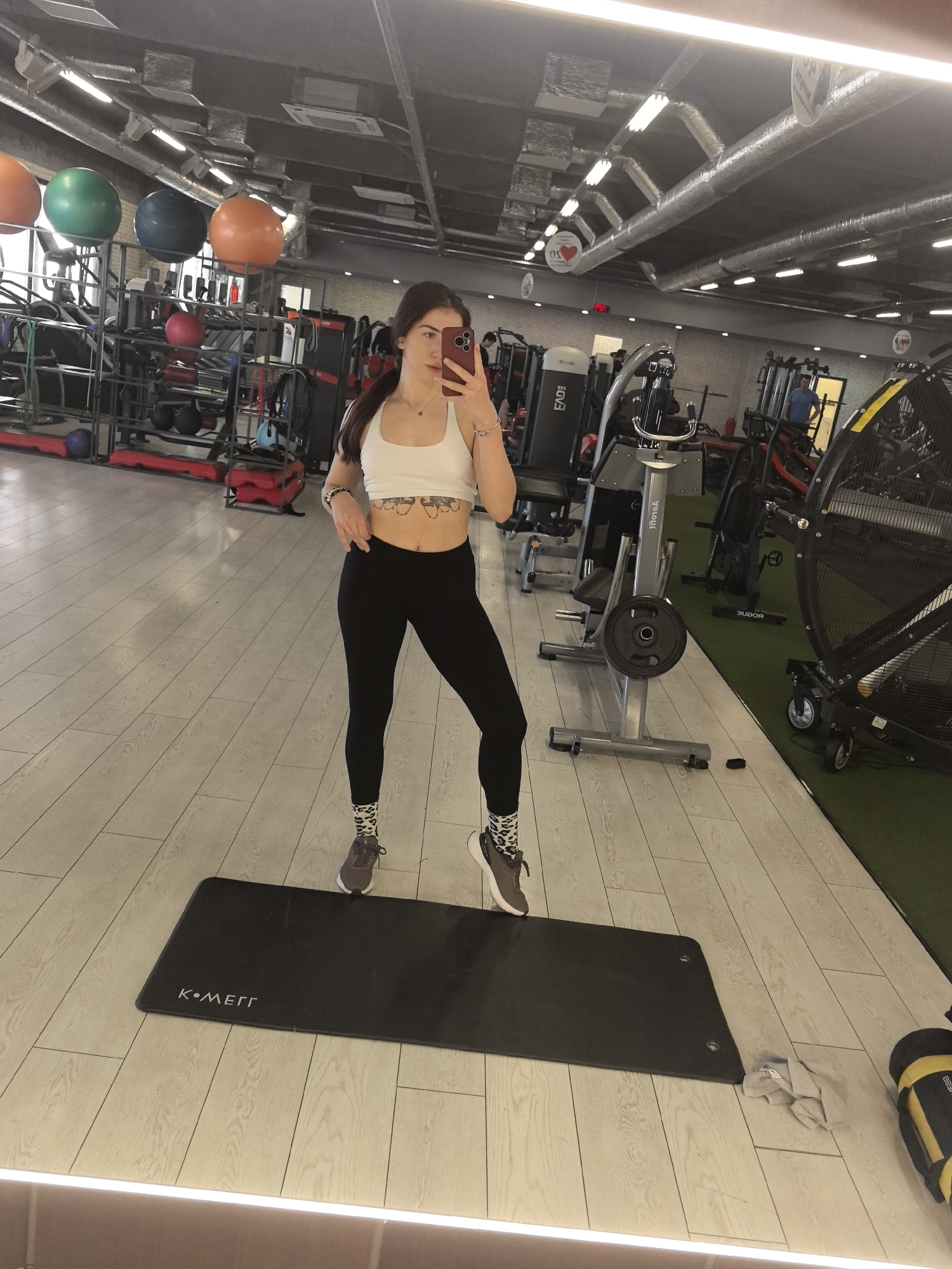 A woman with dark hair is standing on a black yoga mat in a gym, taking a selfie with a smartphone. She is wearing a white sports bra and black leggings with patterned socks and athletic shoes. The gym has a variety of exercise equipment, including weight machines, treadmills, and exercise balls. The floor is light wood, and the ceiling has exposed pipes and ventilation systems. The gym is located in Stara Zagora, Bulgaria.