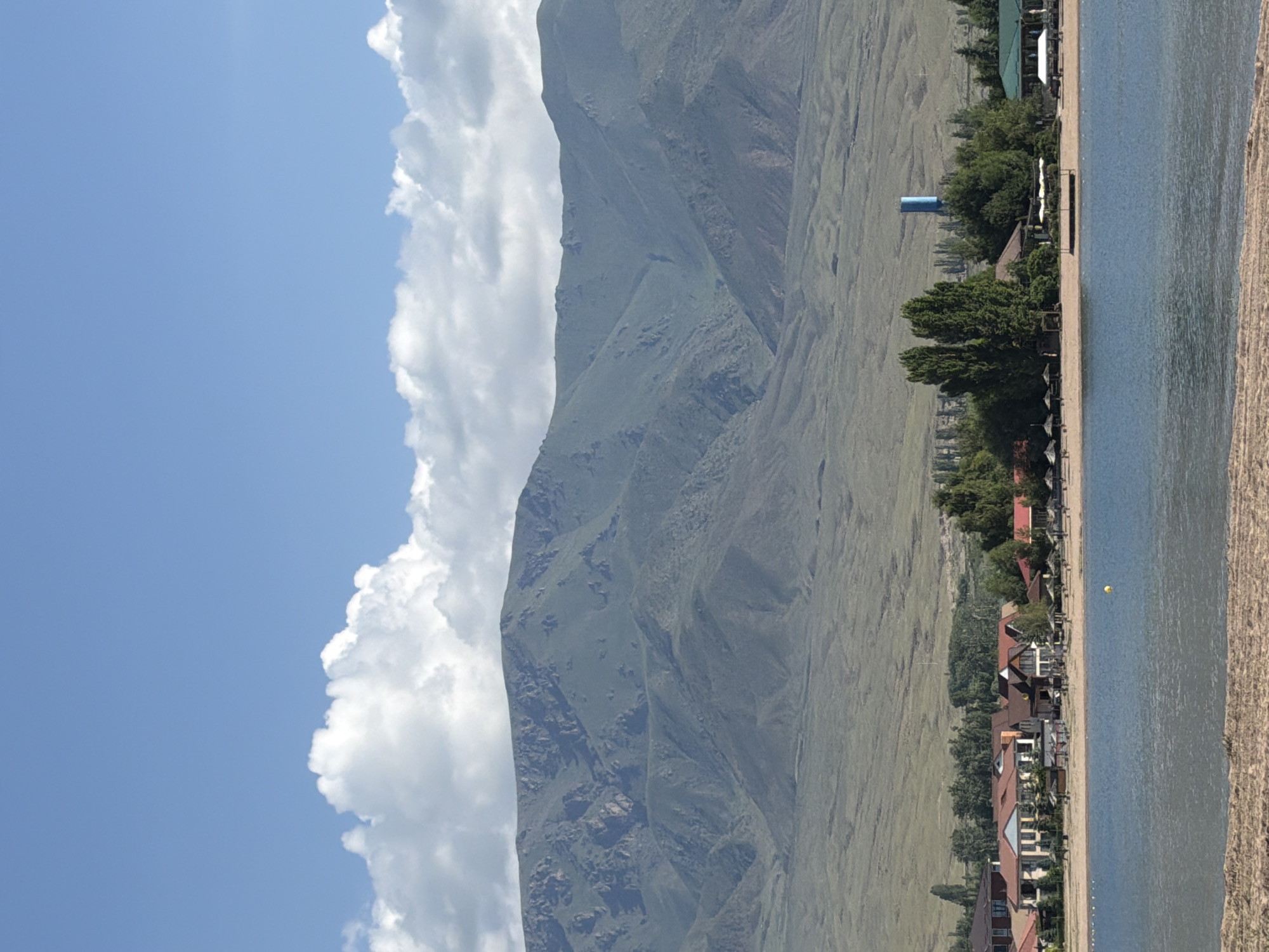 A wide-angle, eye-level, outdoor, daytime shot captures a serene lakeside scene with mountains in the background. The foreground is dominated by a sandy beach and the calm, blue water of a lake. Along the shoreline, a stretch of resort-like buildings with red-tiled roofs and beige facades are visible, interspersed with lush green trees. Some of these buildings appear to be hotels or restaurants, with umbrellas and seating areas on the beach. Further back, a towering blue water tower stands out against the greenery. The middle ground features rolling, green hills leading up to majestic, rugged mountains. The sky above is a vibrant blue, punctuated by fluffy white clouds. The scene evokes a sense of tranquility and natural beauty.