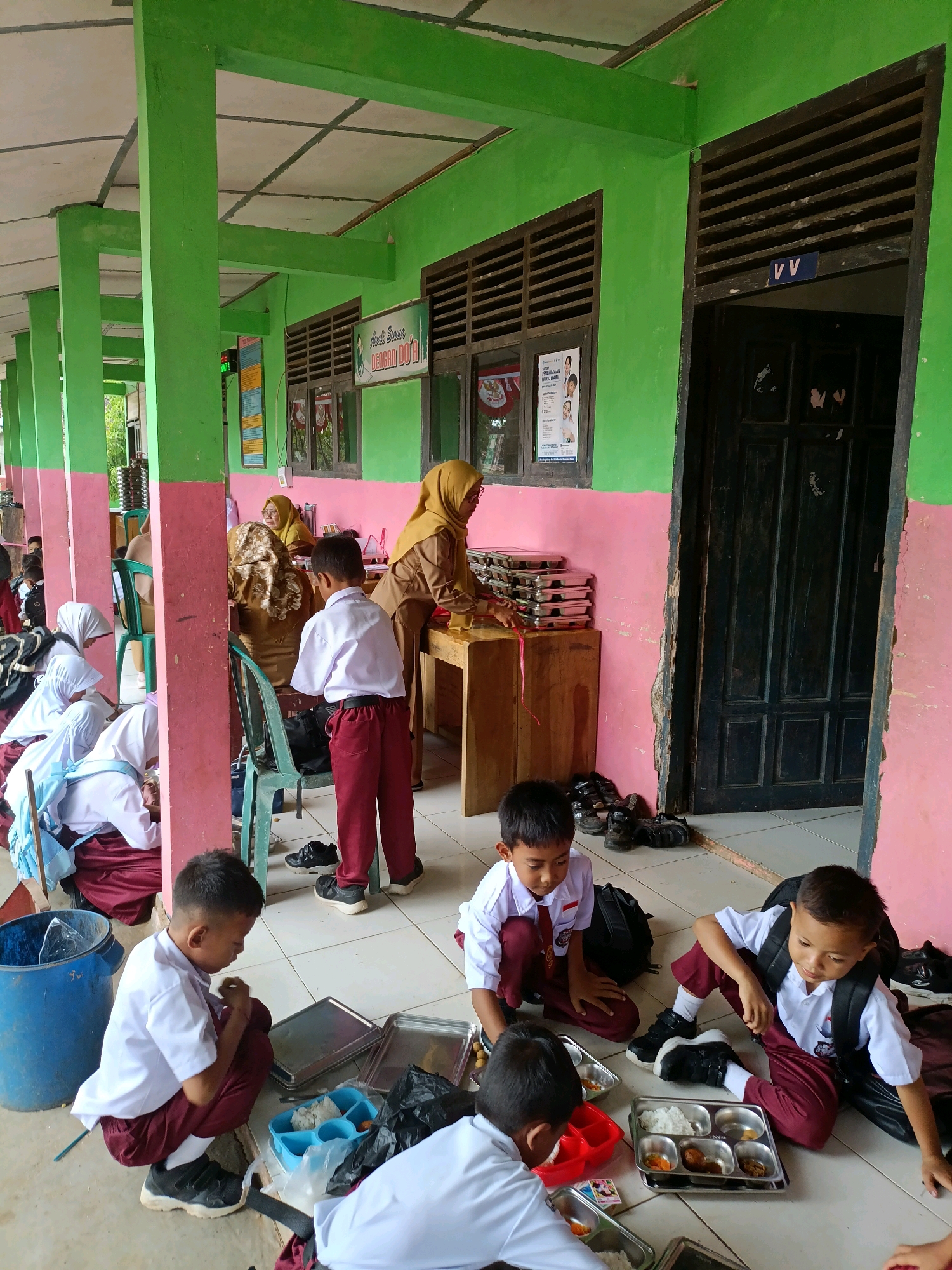 The image depicts an outdoor covered area, likely a school corridor or veranda, in the city of Sumberejo, Indonesia. Several young students in white shirts and maroon pants are present. Some are seated on the floor, eating from metal trays containing rice and other food items. Others are standing or sitting on chairs. Two adult women, dressed in headscarves and school uniforms, are visible in the background, near a wooden table laden with stacked metal containers. The environment is characterized by brightly painted green pillars and pink walls, with a tiled floor. A large blue trash can and various backpacks are also visible. The scene appears to be a school lunch service.