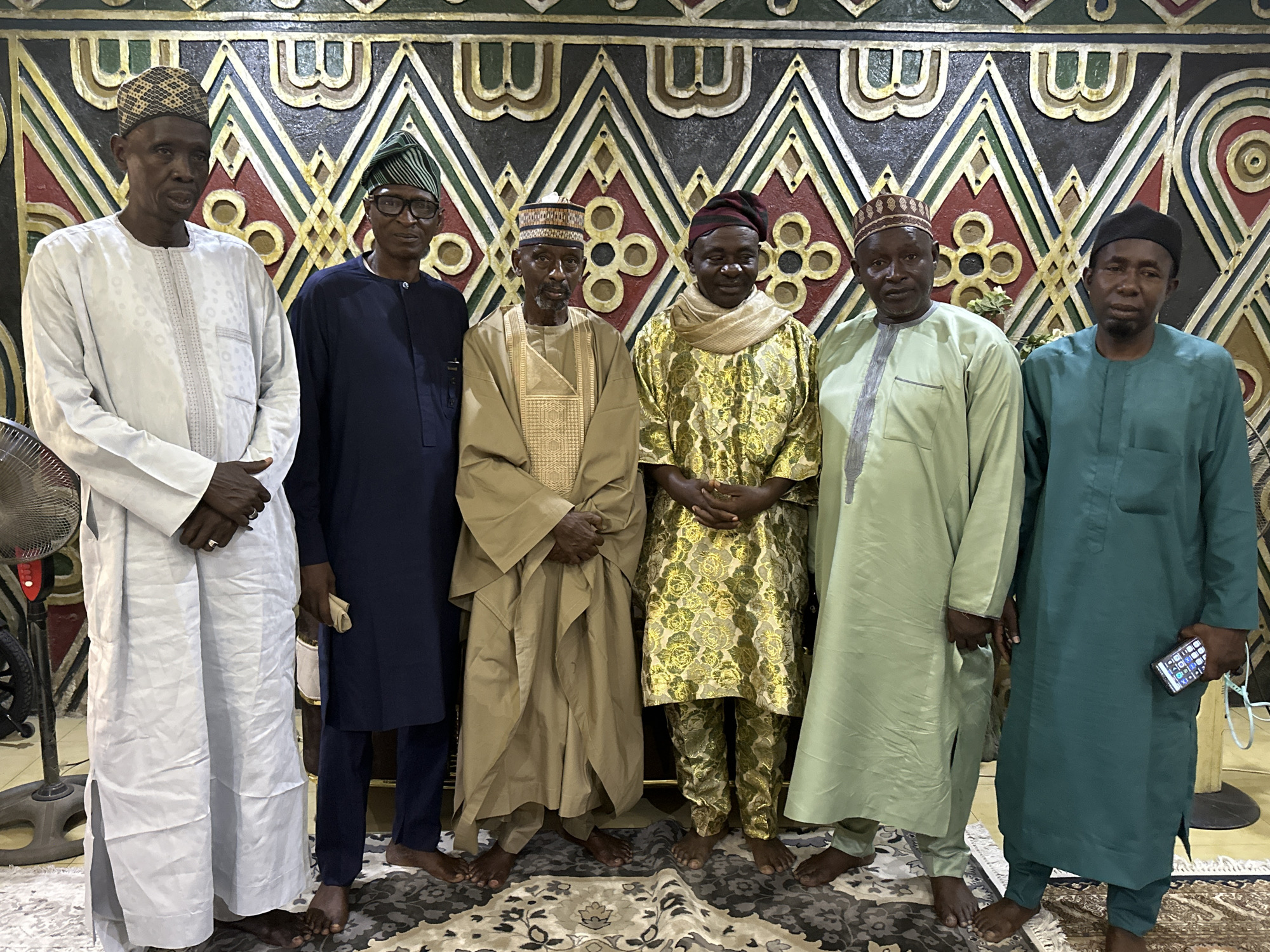A group of six men are standing in a row indoors, posing for a photo. They are dressed in traditional attire, with each man wearing a different style and color of robes and hats. The man on the left is wearing a long white robe and a patterned hat. Next to him is a man in a dark blue robe with a green striped hat. The third man is wearing a tan robe and a patterned hat. The fourth man is wearing a gold patterned robe and scarf with a reddish-brown hat. The fifth man is wearing a light green robe and a patterned hat. The man on the right is wearing a dark green robe and a black hat, and he is holding a smartphone in his right hand. All the men are barefoot. The background consists of a wall with ornate, geometric patterns in black, gold, red, and white. The floor is covered with a patterned rug. To the left of the frame, a partially visible fan can be seen. The location is Unknown, Unknown.