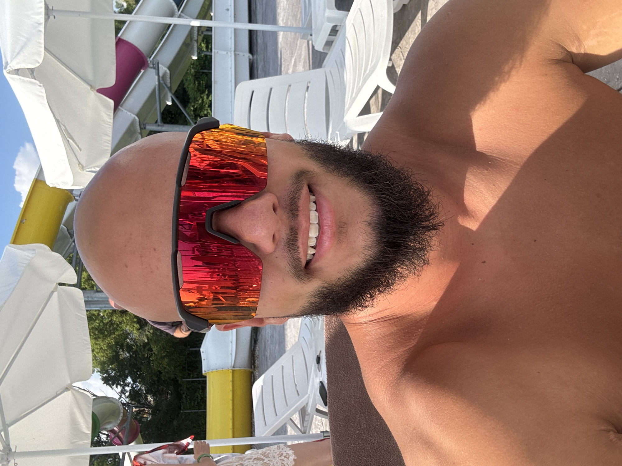 A close-up, shoulder-up selfie of a bald, bearded man wearing mirrored sunglasses with red and orange lenses. He is outdoors, possibly at a water park, with white umbrellas and water slides visible in the background. The man is shirtless, and the lighting suggests it is a sunny day. The location is identified as Ivanyane, Bulgaria.