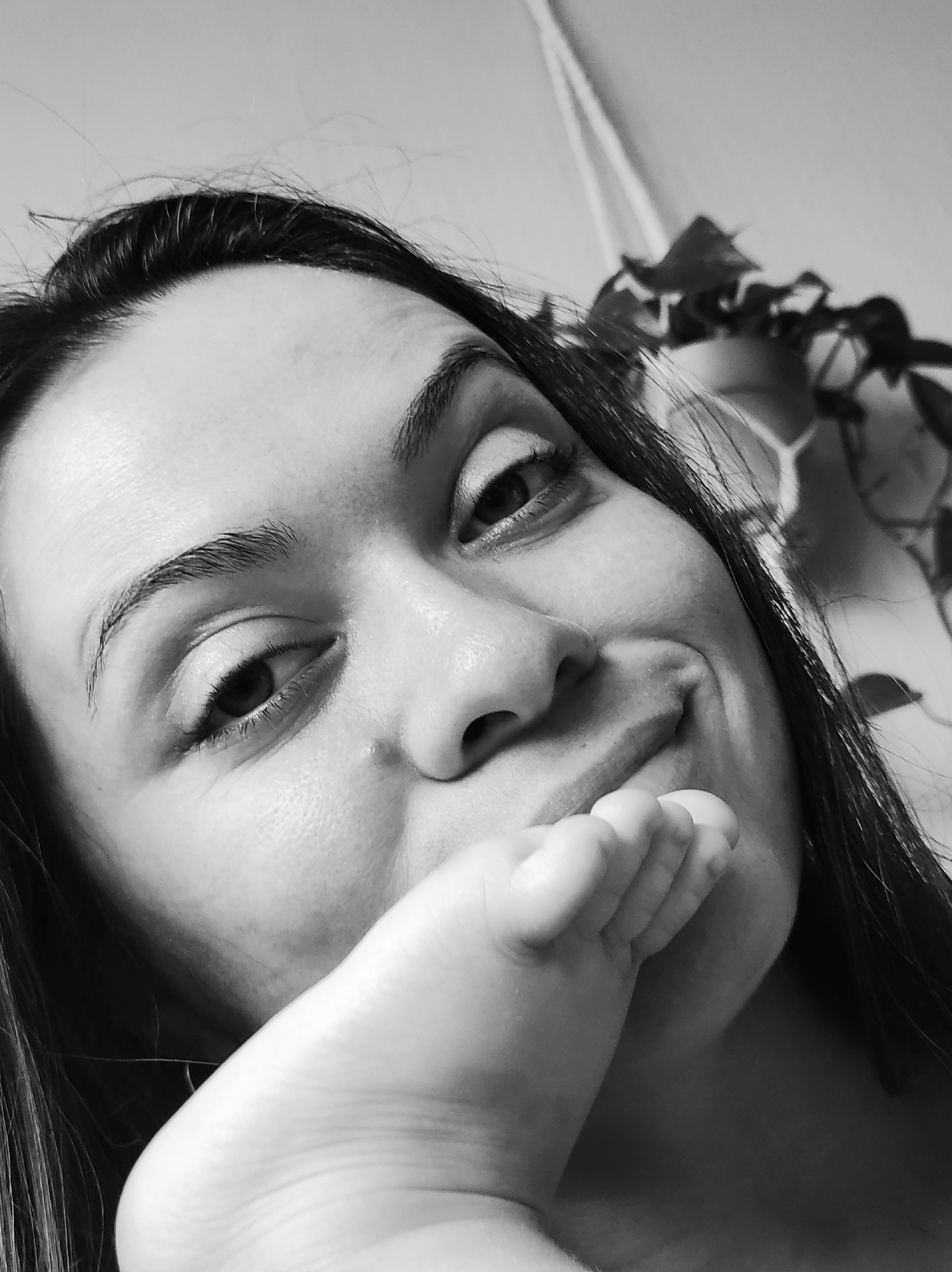 The image is a close-up, black and white, low-angle selfie of a woman. Her face fills most of the frame, with her head tilted and her gaze directed slightly upwards. A foot is held up to her mouth, with its toes resting against her lips and cheek. She has dark hair, with strands falling around her face. Her expression is a playful smile, with dimples visible on her cheeks. The background is softly blurred, showing what appears to be a plant with leaves to the right of her head and a plain surface behind it, possibly a wall or curtain. There is no information available about the location of Chaikovsky, Russia within this image.